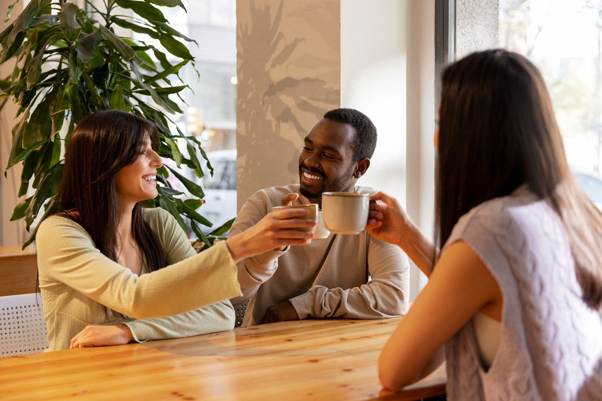 Three people clink their mugs while sitting together at a wooden table in a cafe with a large plant nearby.