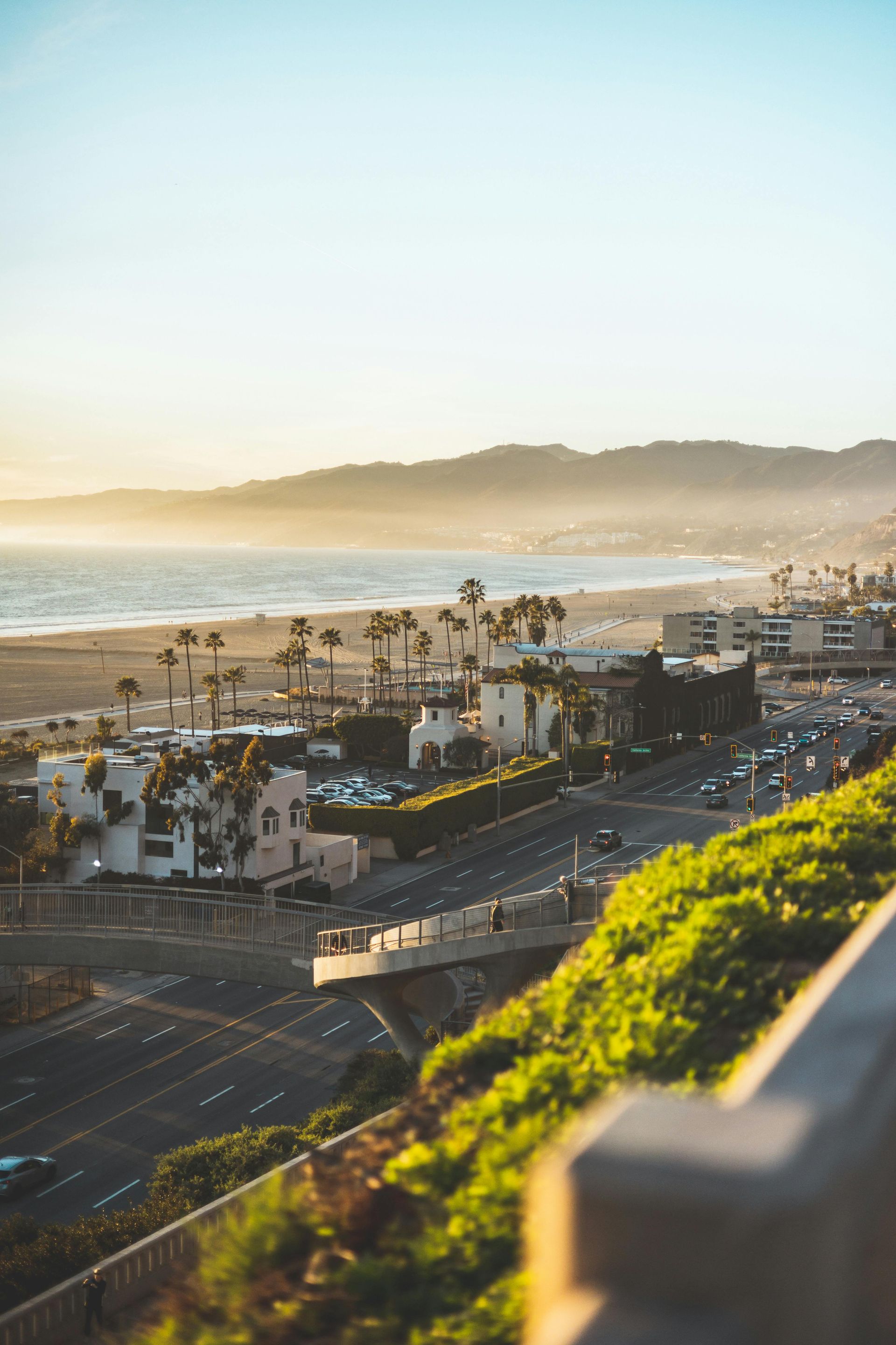 Golden hour sunlight shines over a beach, coastal town, and highway, framed by green hillside foliage in the foreground.