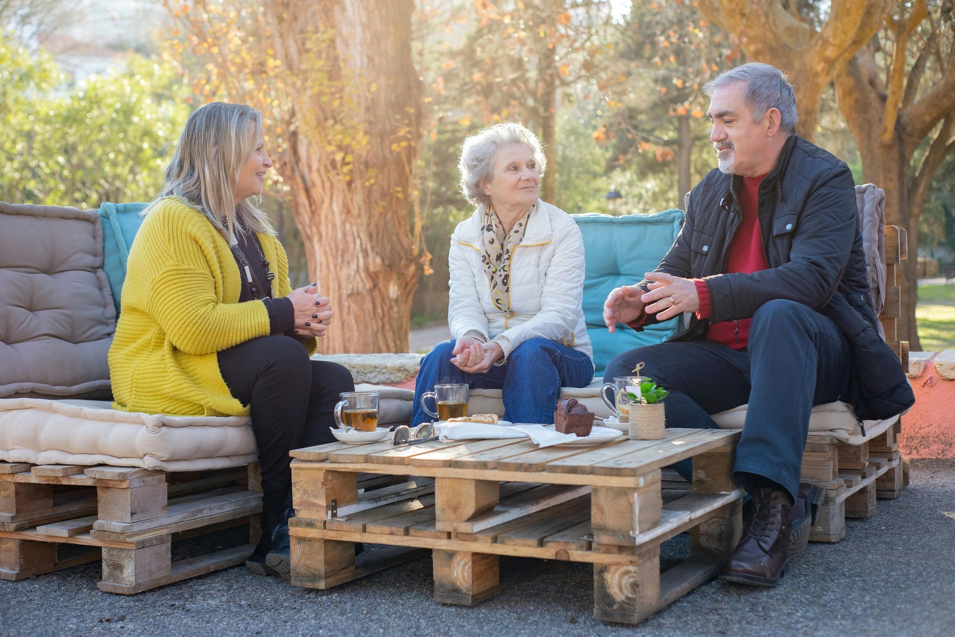 Three people sit on pallet furniture outdoors, chatting over drinks and snacks in a bright, sunlit park setting.