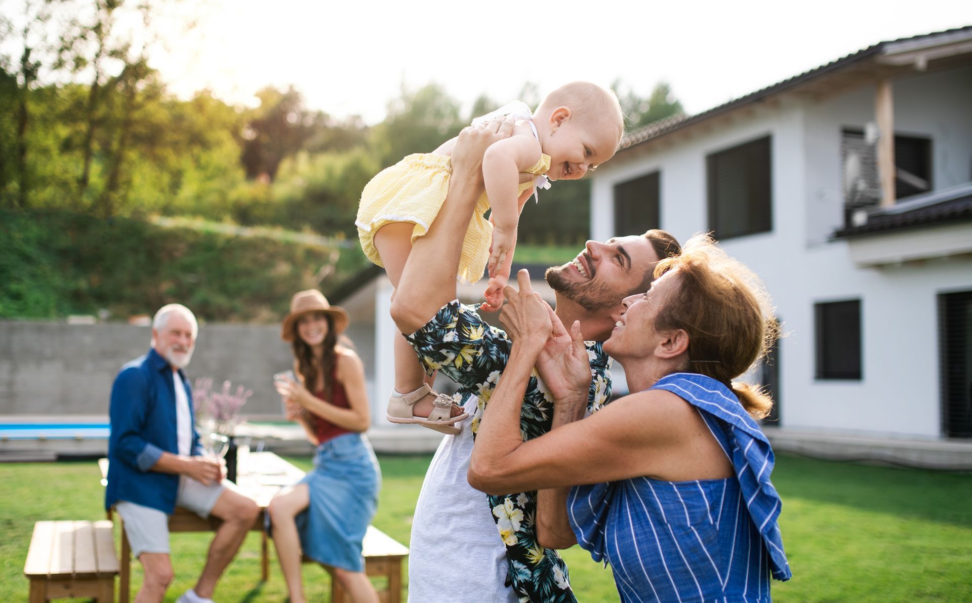 A family gathers in a sunny backyard as an adult lifts a toddler in the air while others smile and socialize.