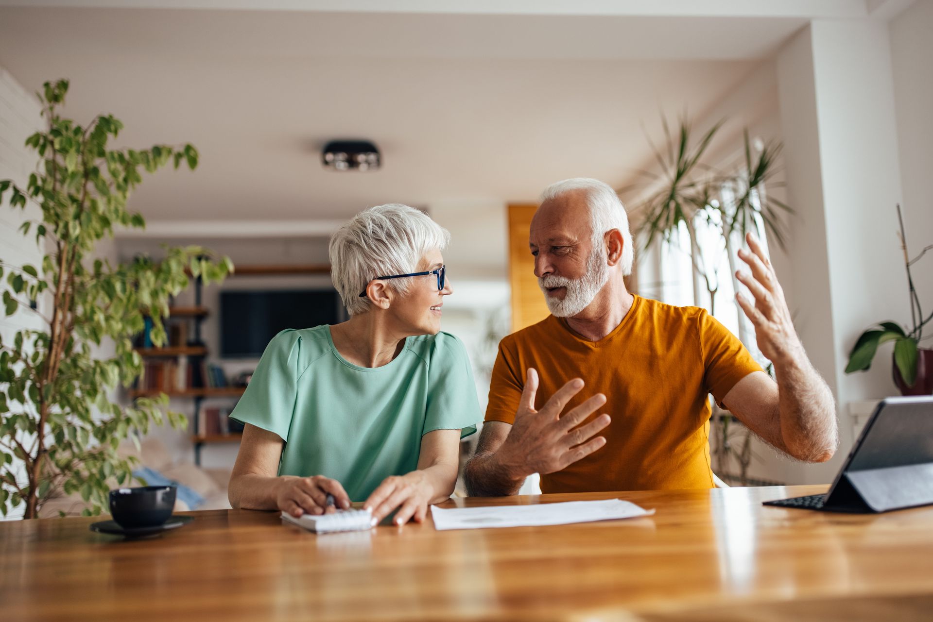 Two people sitting at a wooden table in a bright room, discussing documents and a tablet computer.
