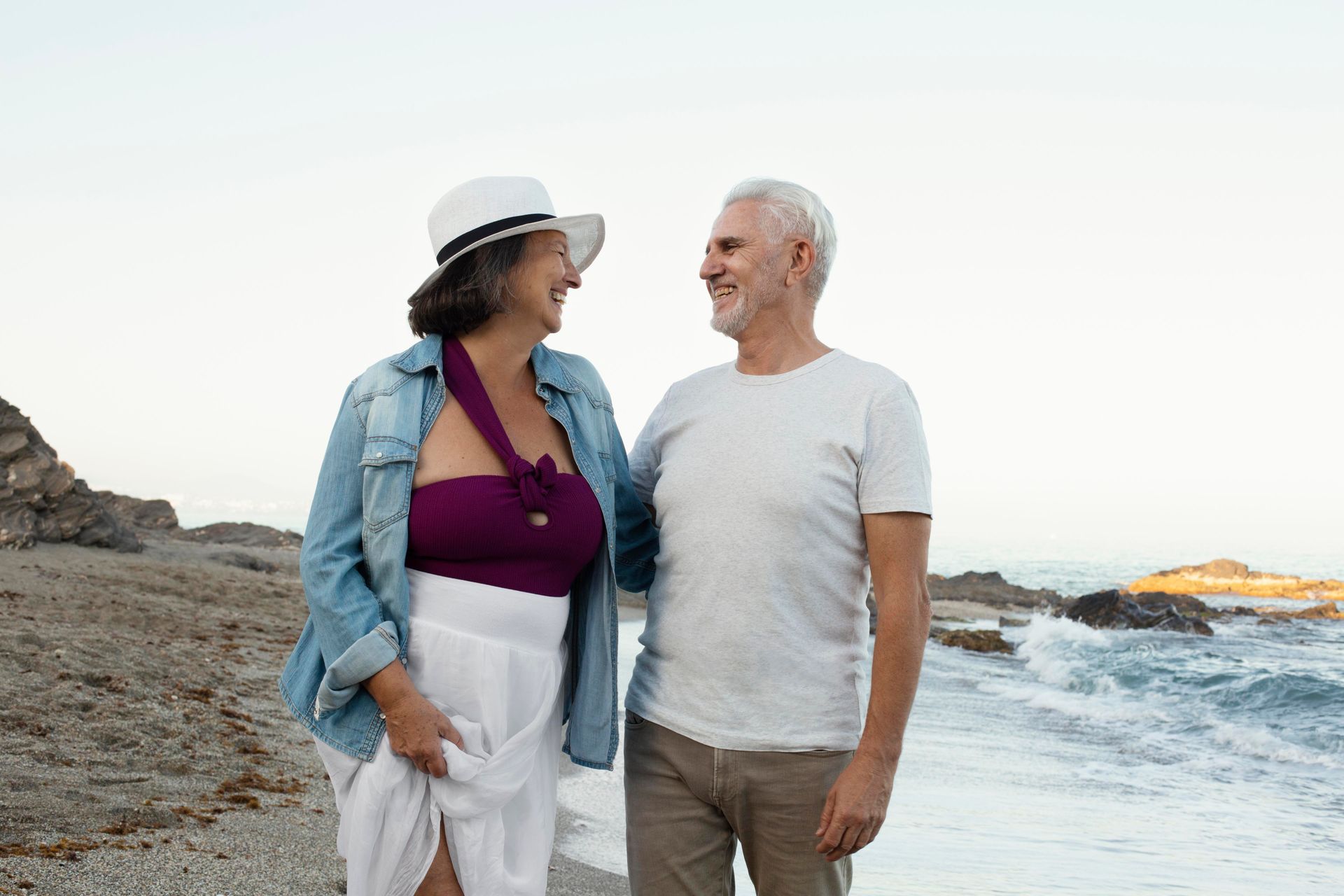 An older couple smiling at each other while walking along a rocky, overcast beach near the shoreline.