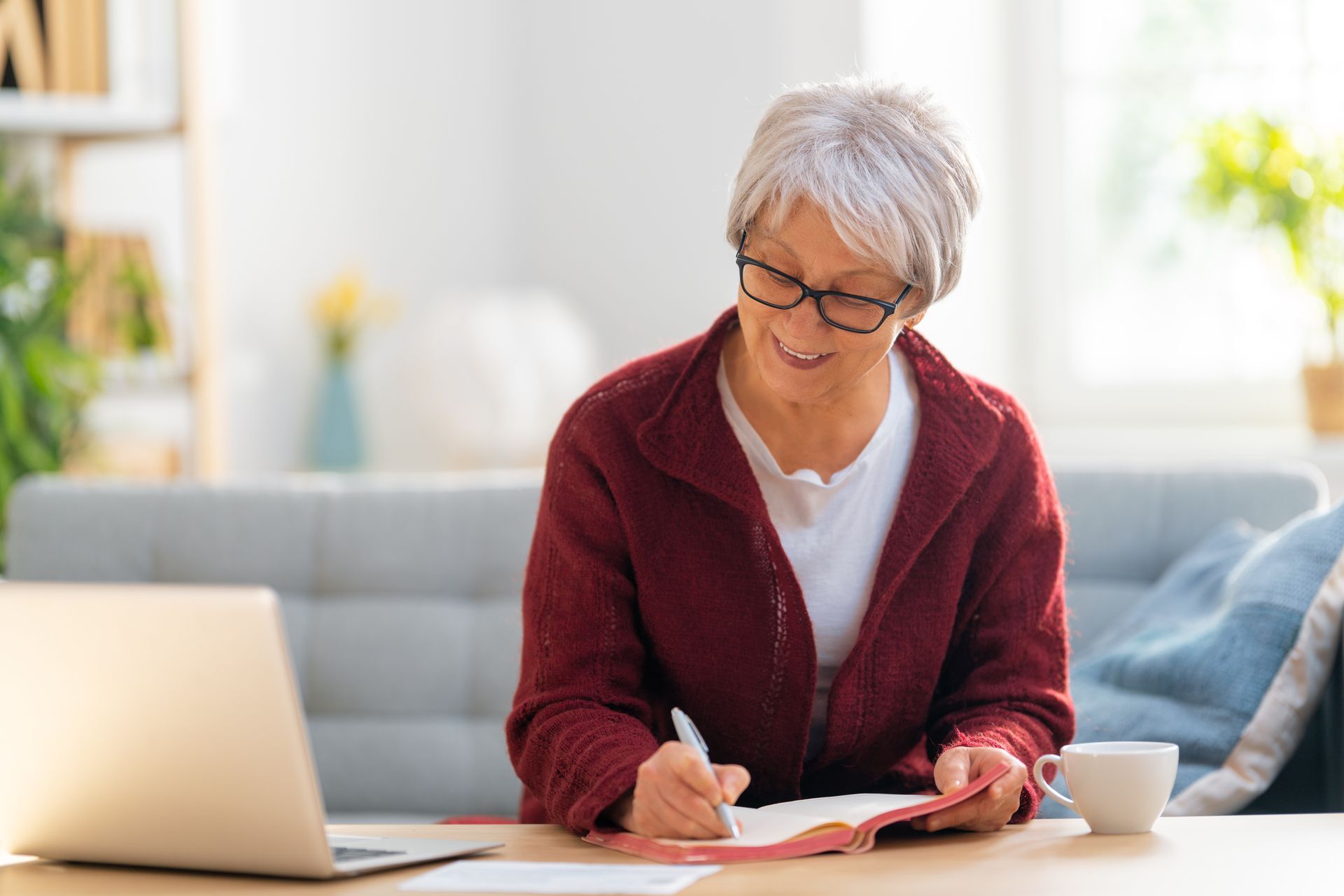 A person with gray hair wearing glasses and a red cardigan writes in a notebook while working at a laptop in a bright room.