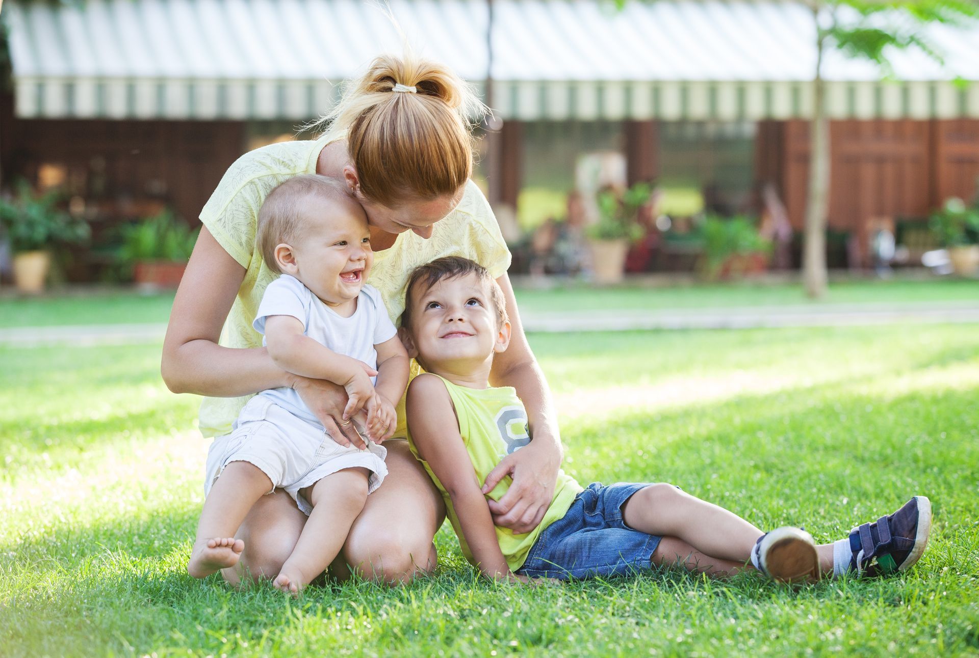 A person sitting on green grass outdoors, holding a laughing toddler and hugging a young child looking up at them.