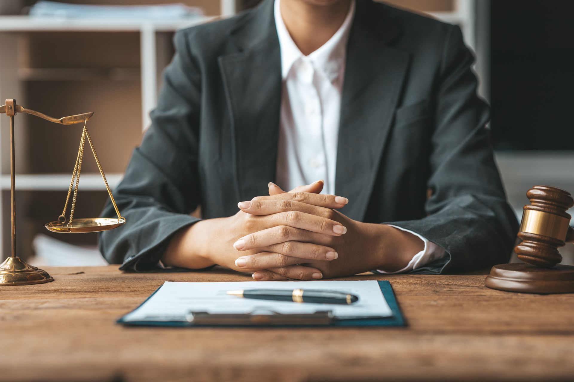 Person in suit with folded hands, legal documents, scale, and gavel on a desk.