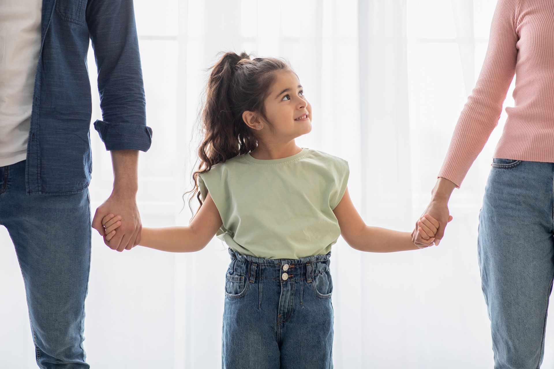 Girl holding hands with two adults, looking upward, inside near a window.