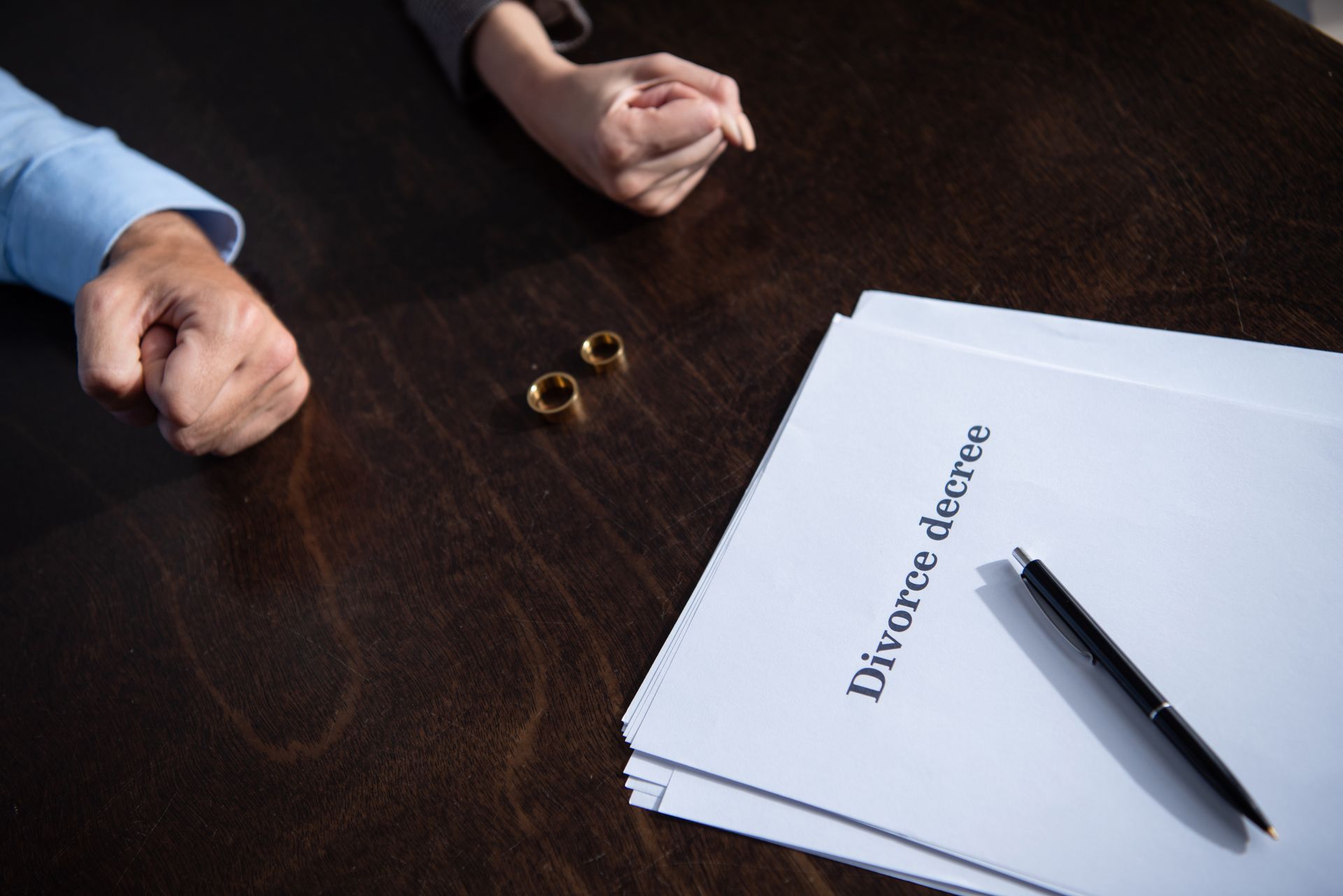 Hands clench near wedding rings and divorce decree documents on a wooden table.