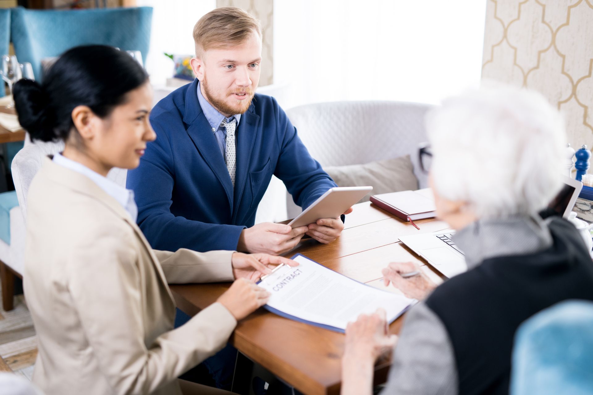 Three people at a table reviewing documents. A man in a suit holds a tablet. Two women examine papers.