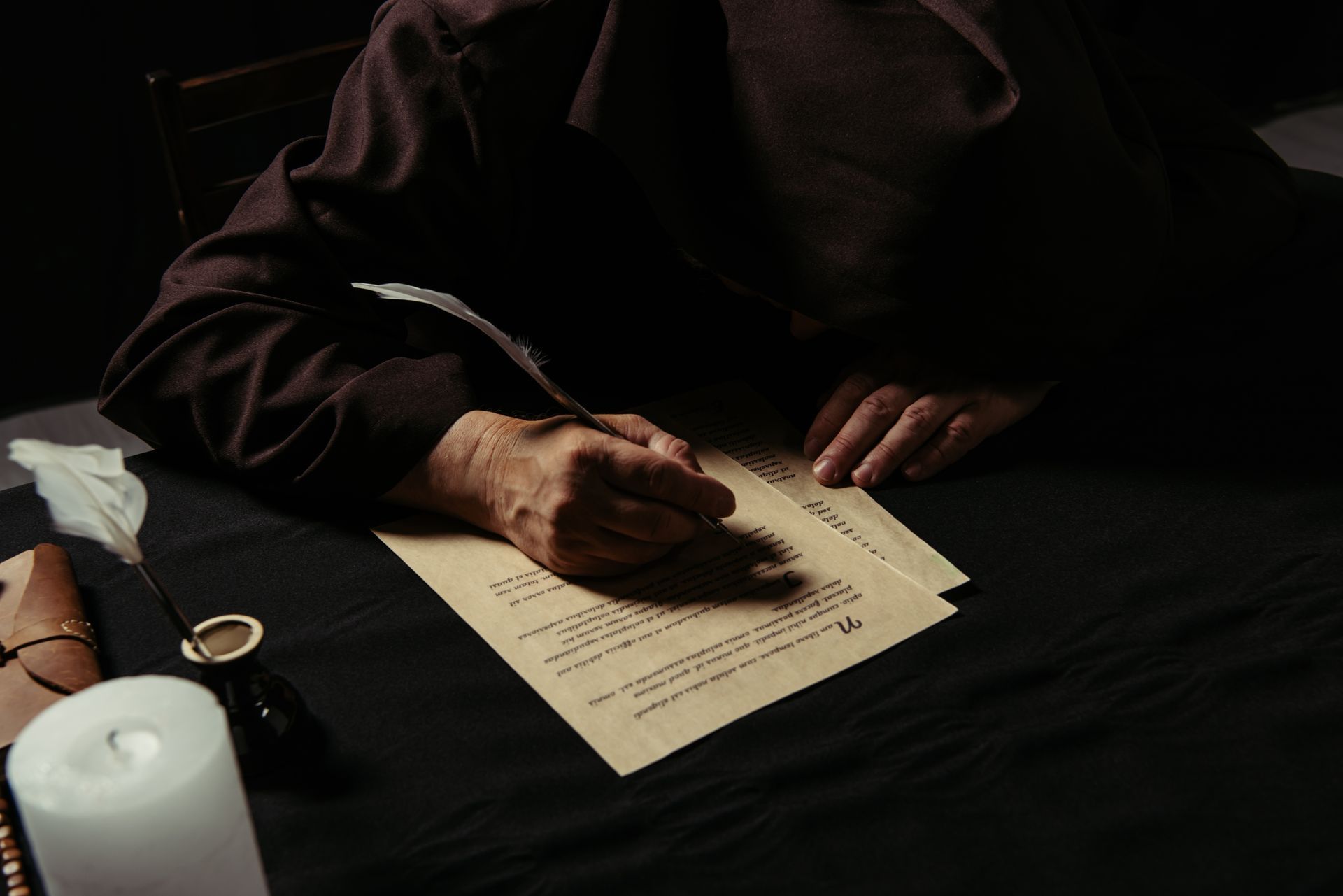 Person in dark robes writing with quill on paper at a desk, lit by candlelight.
