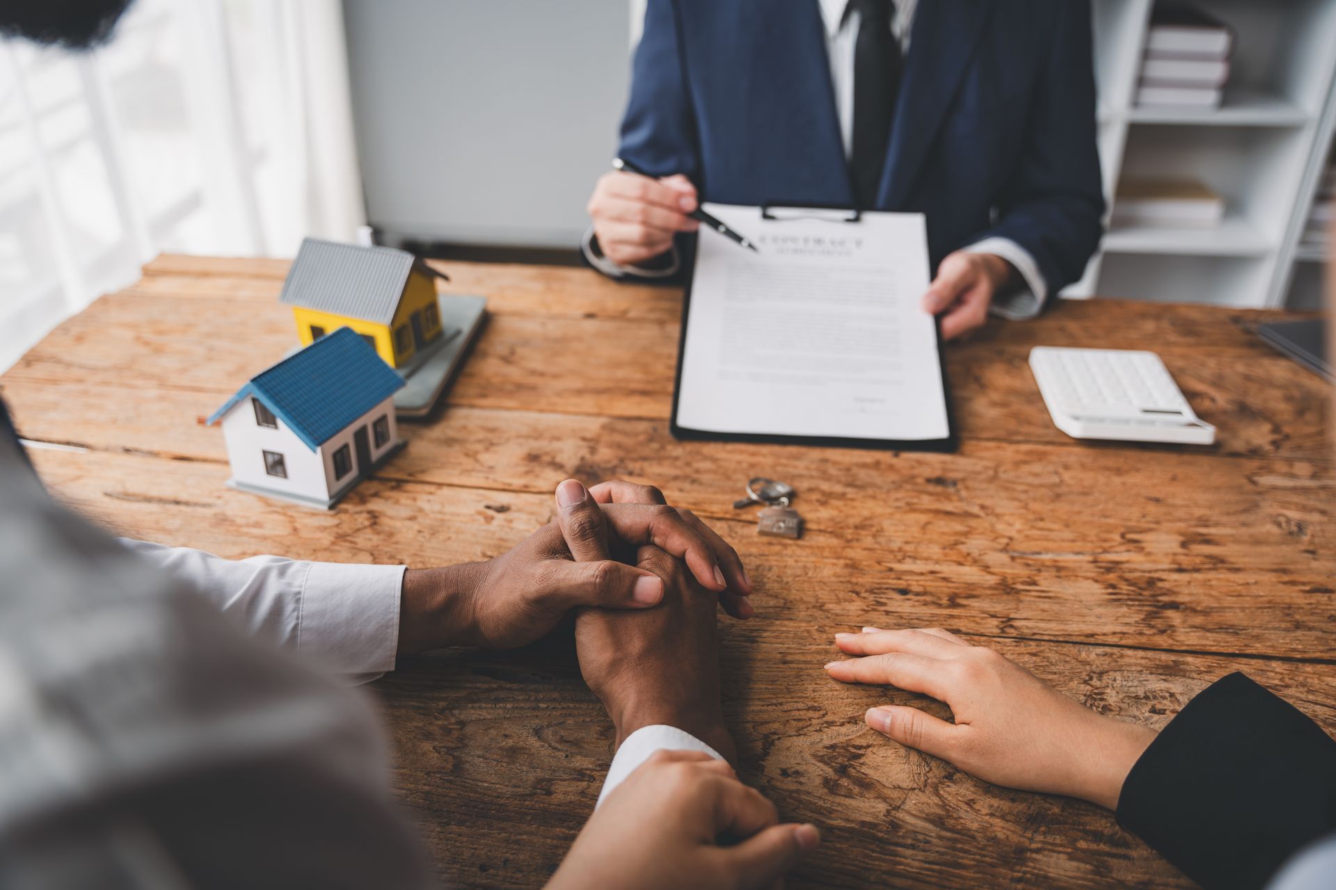 Couple holding hands at a desk, reviewing a document with a real estate agent; model houses sit nearby.