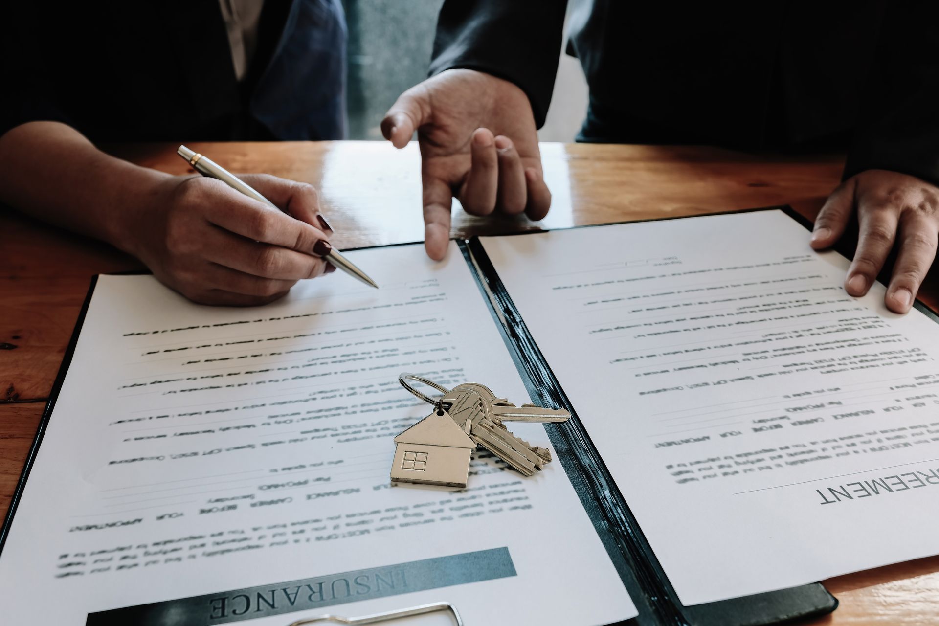 Hands pointing at and signing a document on a table, with keys and a house-shaped charm.