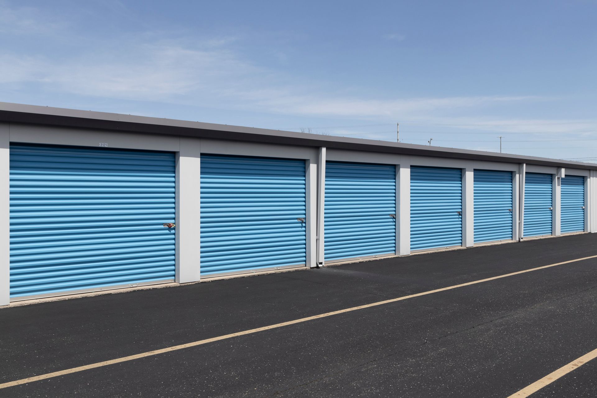 Row of blue storage unit doors with a paved road in front and blue sky. Row of blue storage unit doors with a paved road in front and blue sky.