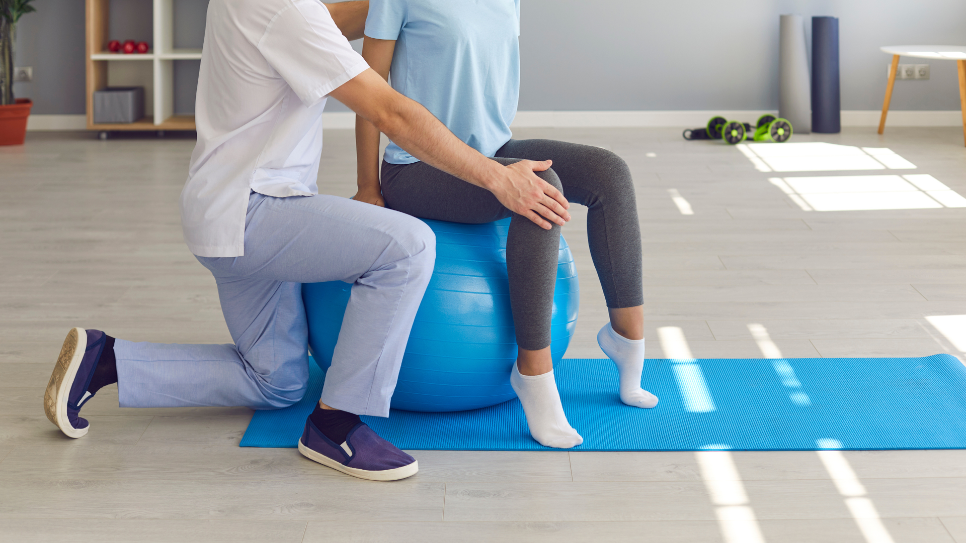 Therapist assists patient with leg exercises while seated on a blue exercise ball. Interior.