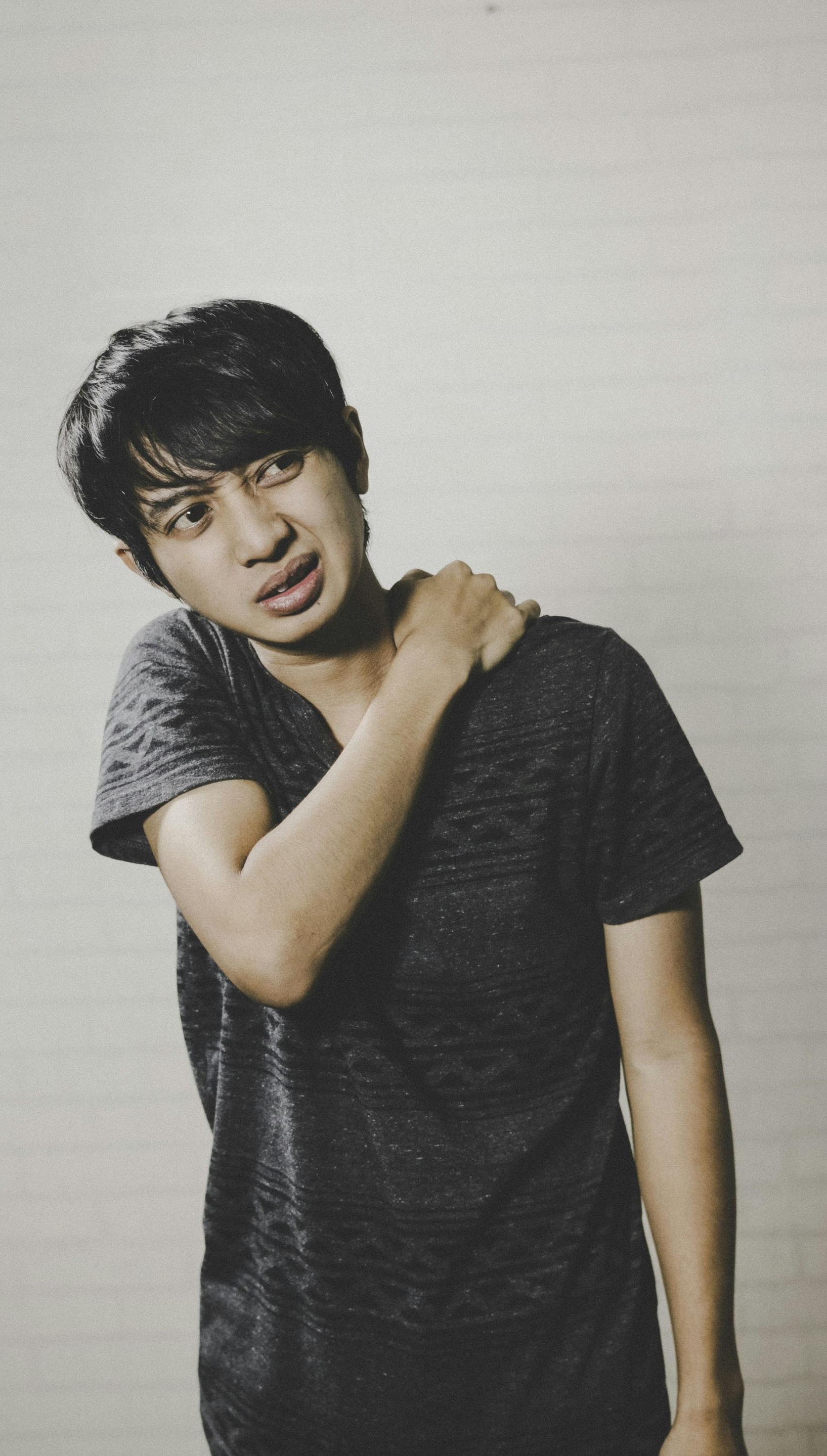 Man grimaces, touching his shoulder. Gray-toned, wearing a dark shirt. Against a neutral background.