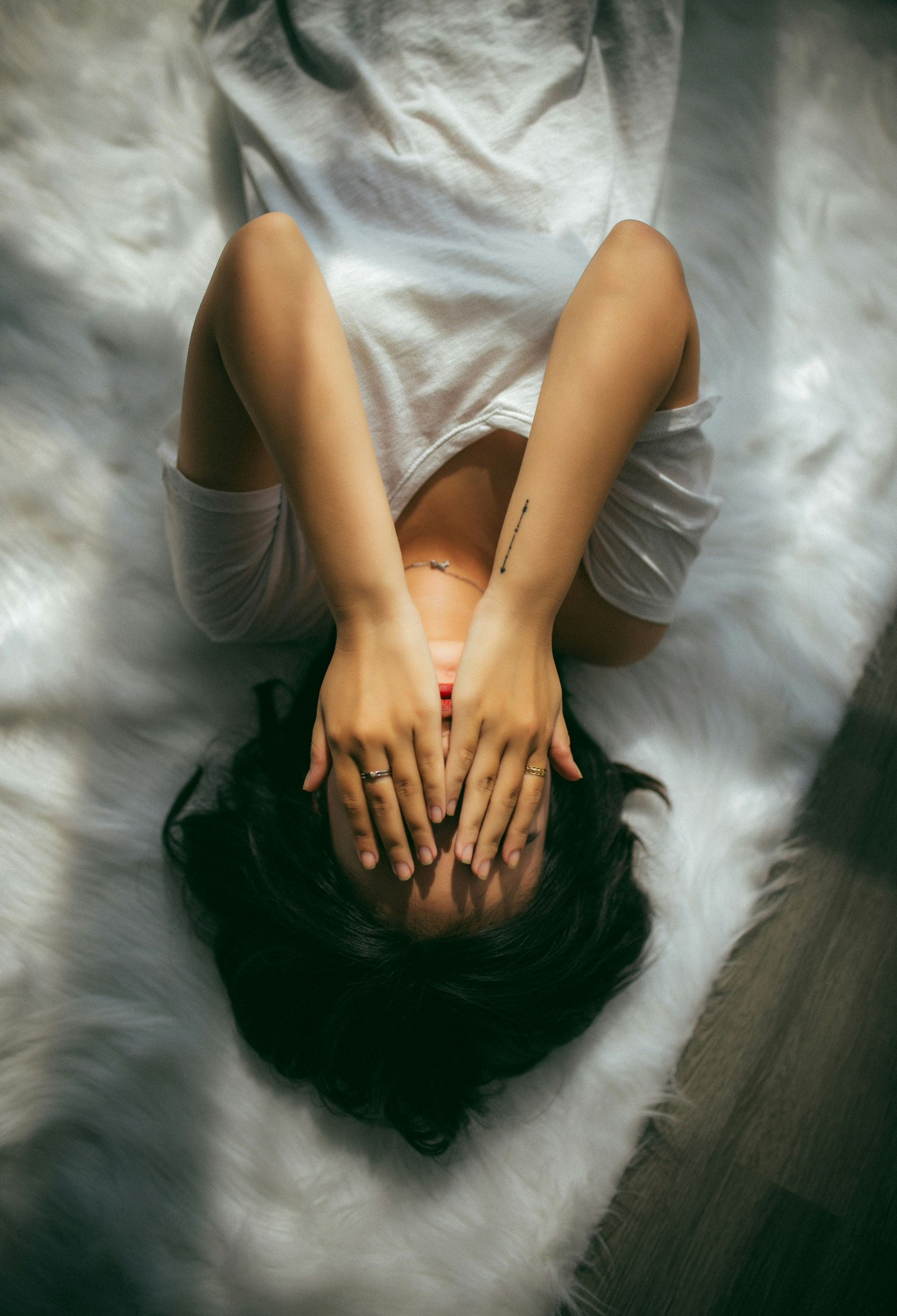 Woman lying on a white fluffy rug, covering her eyes with hands. Sunlight streams in.