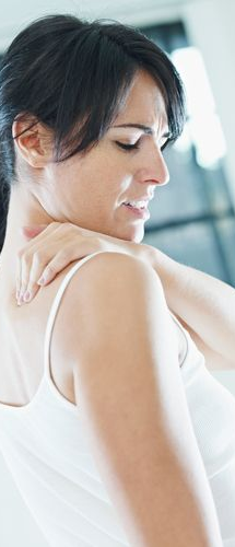 Woman in a white tank top touching her shoulder, appearing to be in pain, with a pained expression.