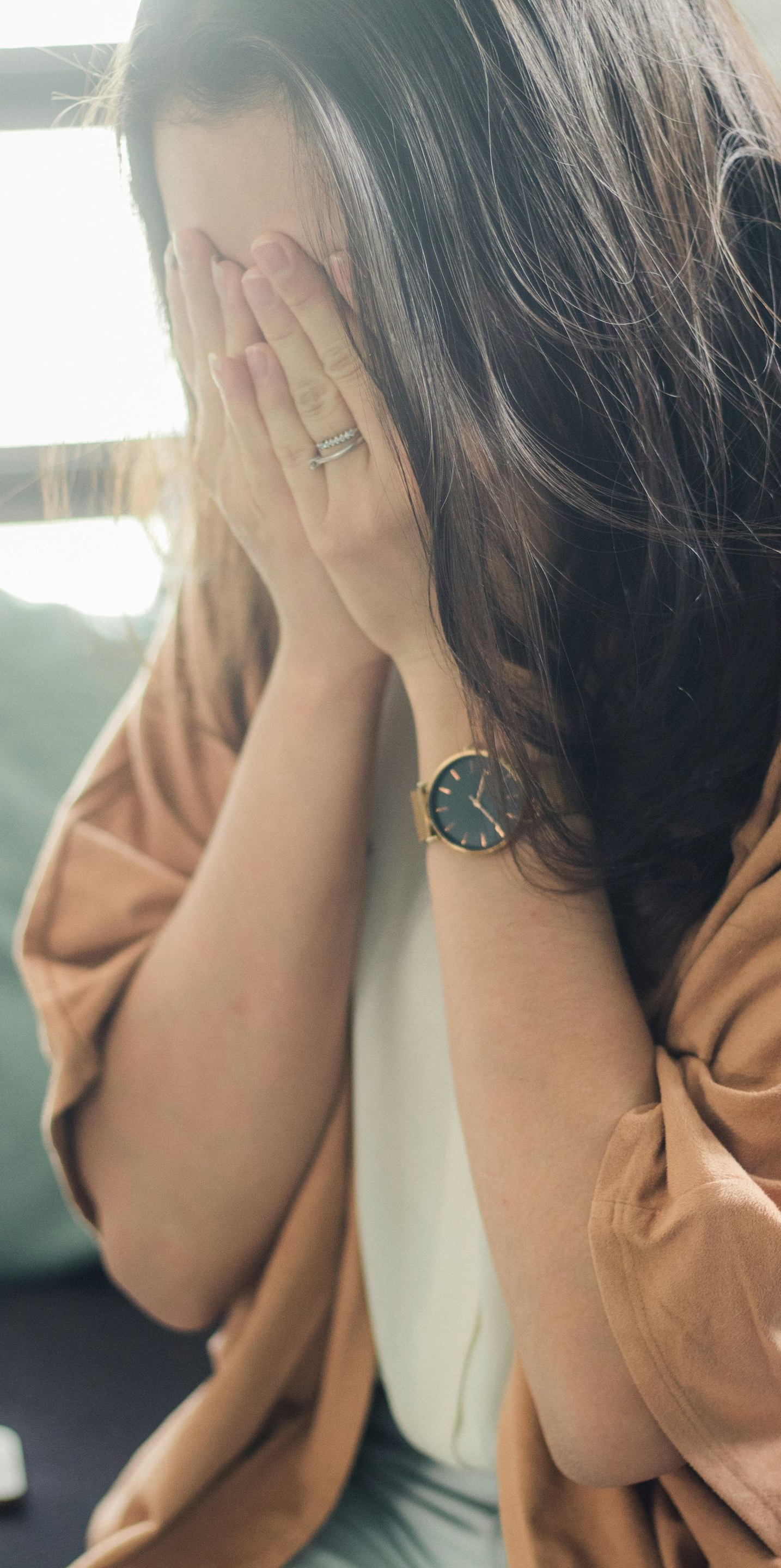 Woman with hands covering face, appearing upset. Wearing watch, sitting indoors.