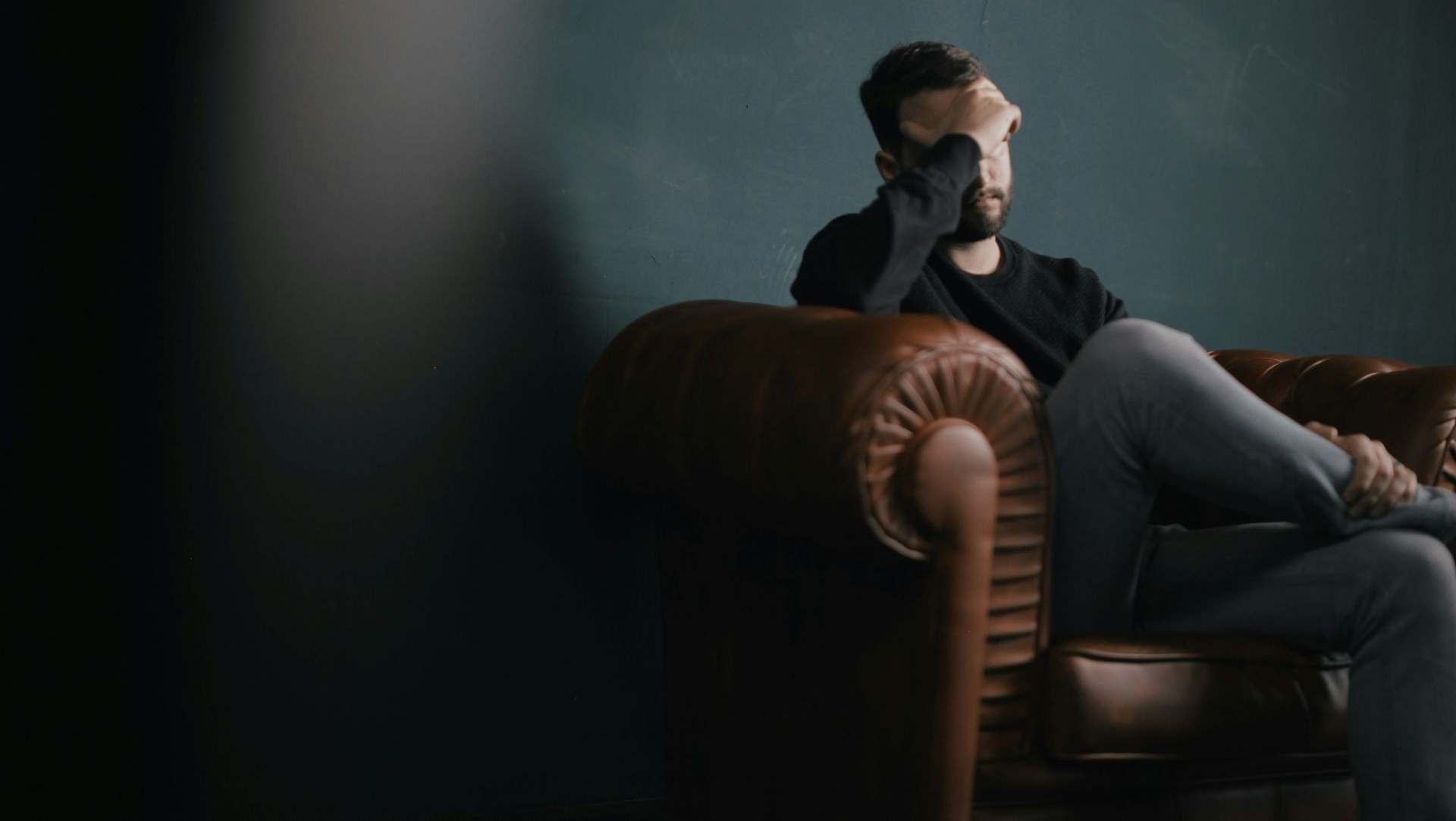 Man sitting on brown couch, hand over face, in a dimly lit room, possibly distressed.