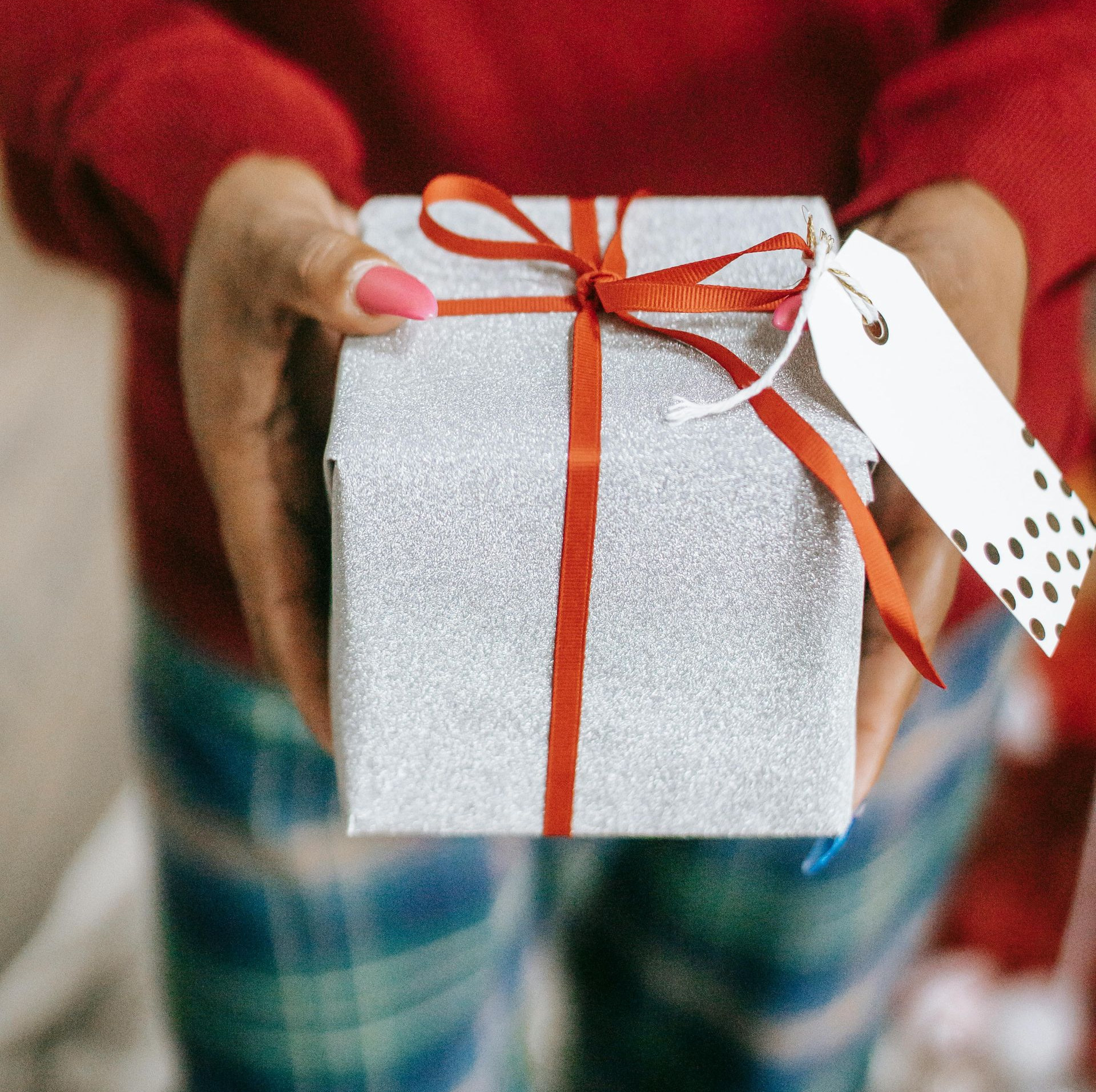Person holding a wrapped silver gift box with a red ribbon and tag.