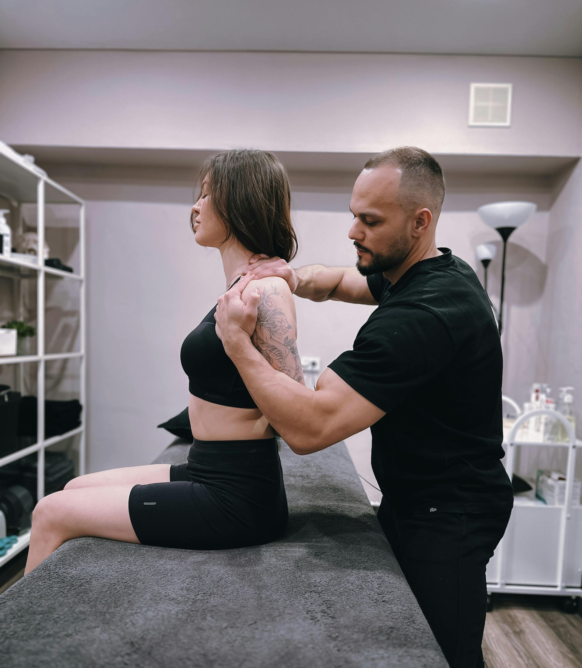 Man assisting woman with shoulder/back mobility on a treatment table. Neutral-toned room.