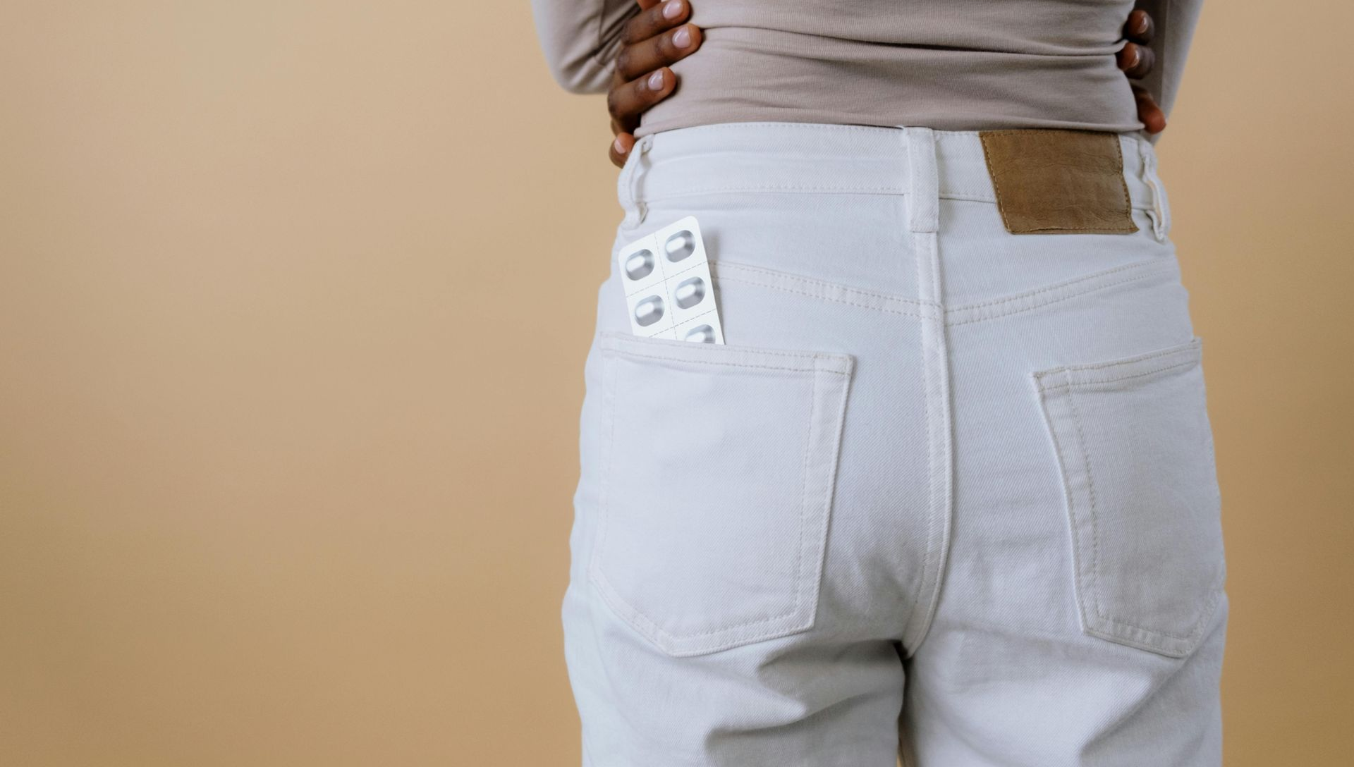 Woman in white jeans with medication in back pocket, arms crossed, neutral background.