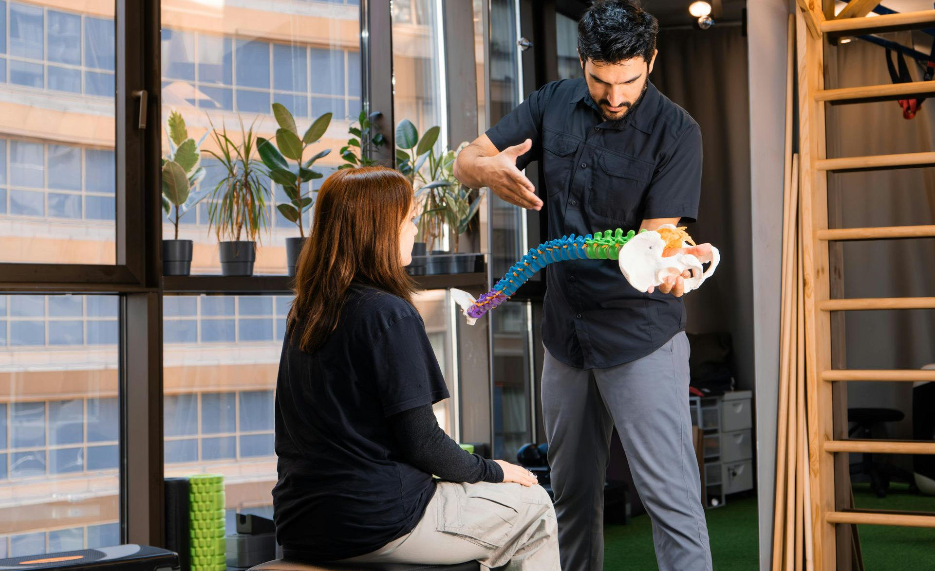 Physiotherapist explaining spine model to patient in a clinic, near a window.