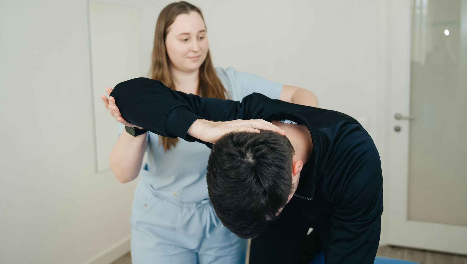Therapist assisting a person with neck movement in a bright room. The person bends forward.