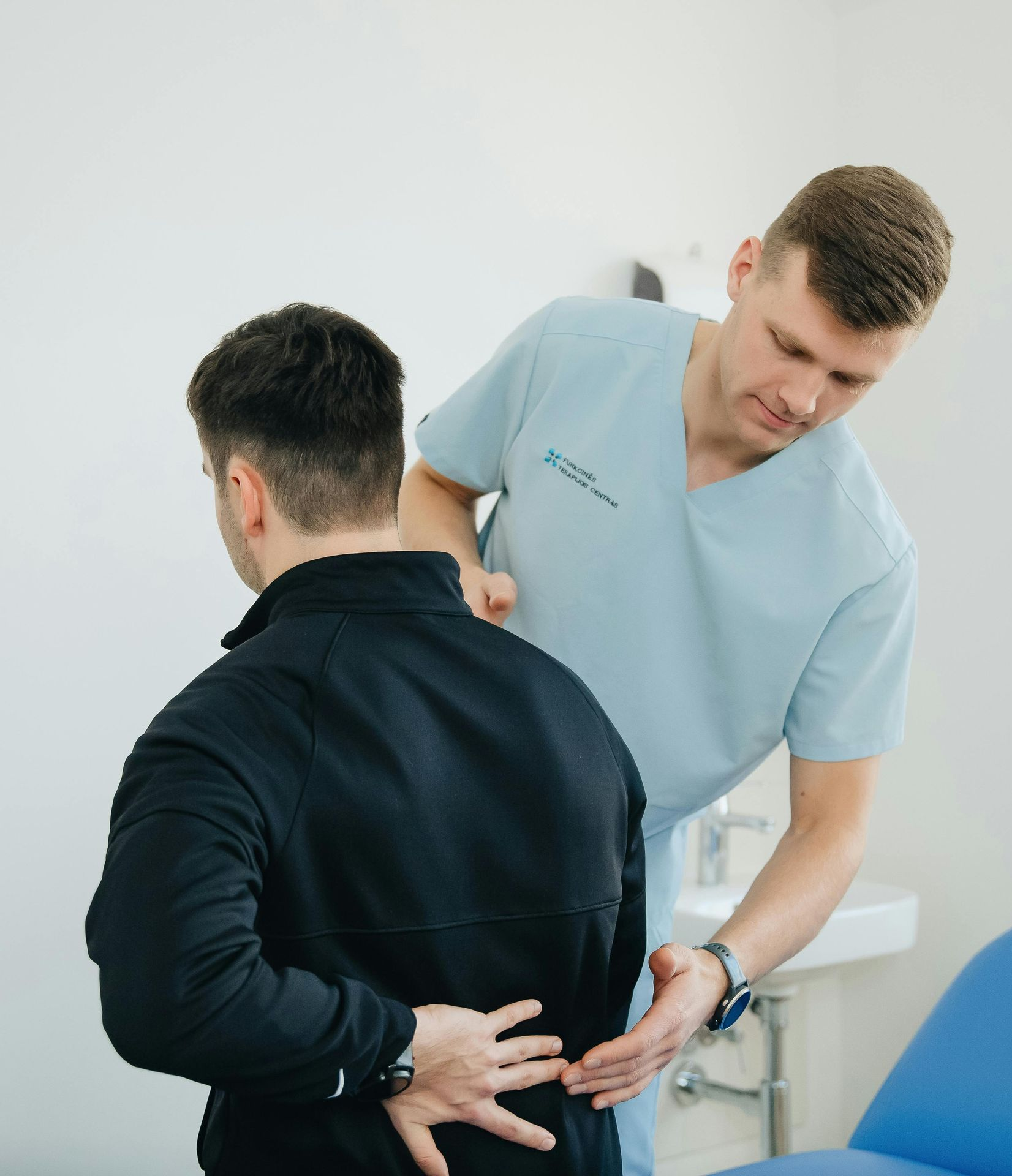 Physician examining patient's lower back in clinic; patient faces away. Blue scrubs, white room.