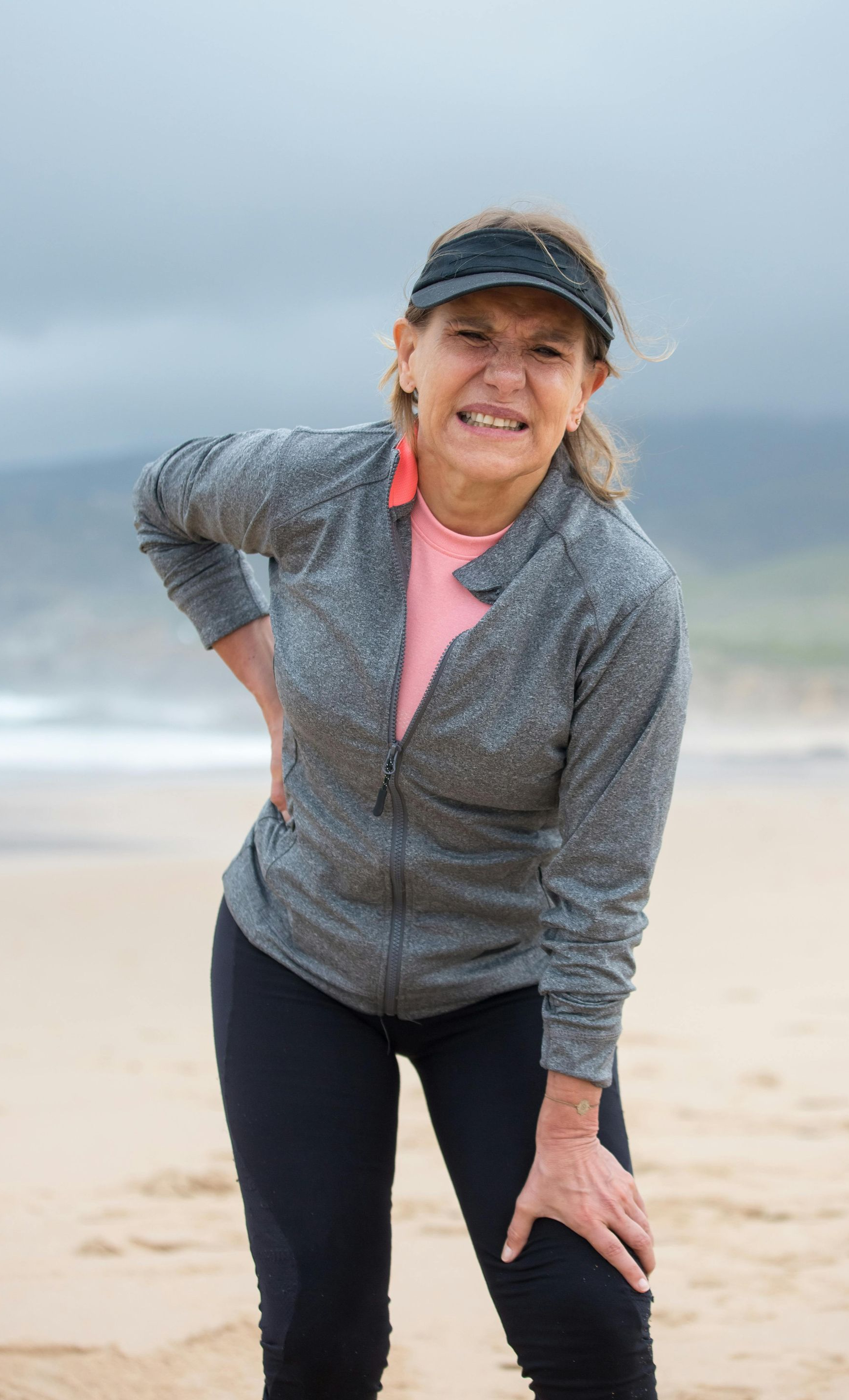Woman on beach in athletic wear, grimacing, holding her knee and lower back. Cloudy day.