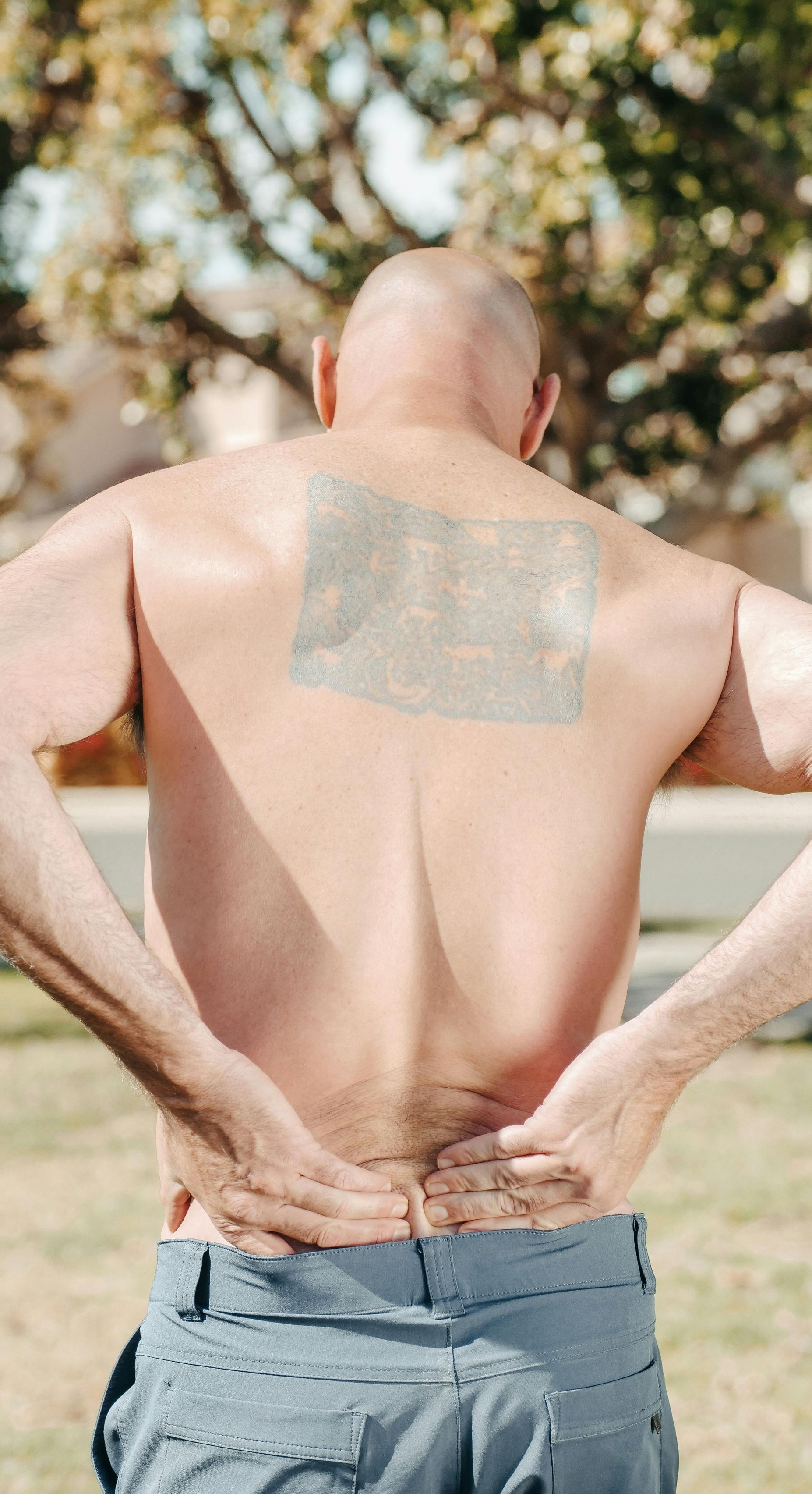 Bald person with hands on lower back, possibly experiencing pain; outdoors.