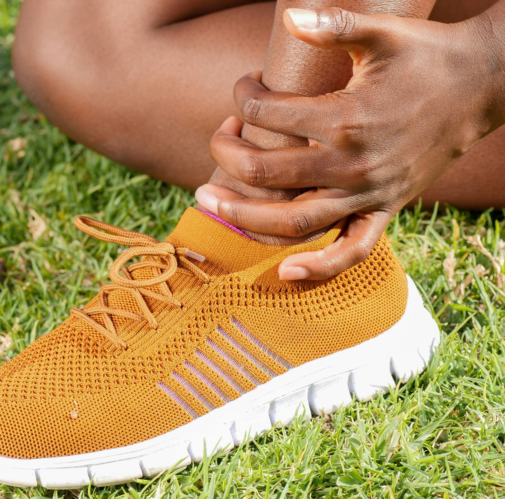 Person holding their ankle, possibly in pain, while wearing a gold sneaker on green grass.