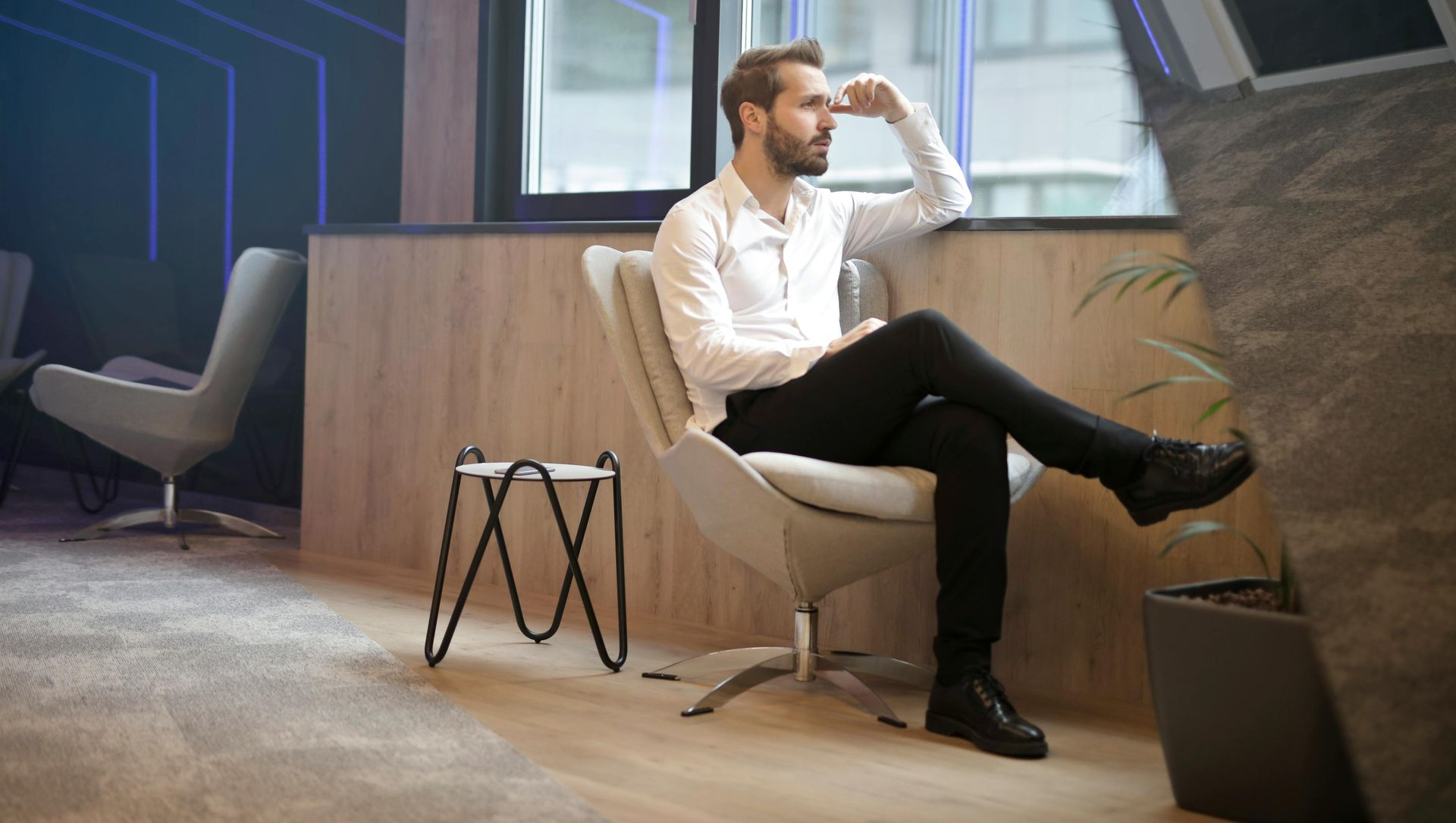 Man sitting, looking out window, resting arm on his forehead. Modern office with chairs, wood paneling.