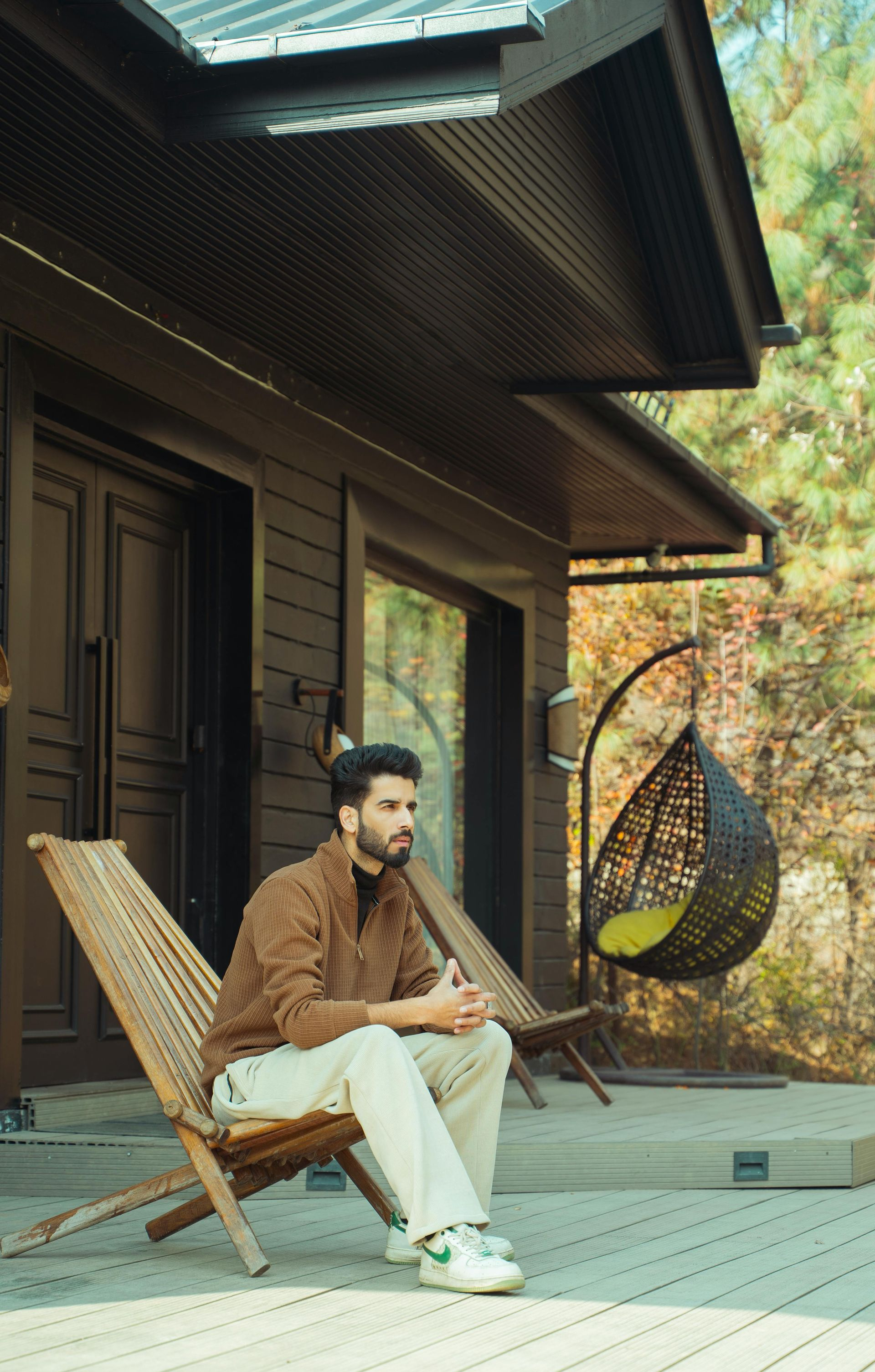 Man sitting on a wooden chair, resting in front of a wooden cabin. He's wearing brown sweater and light pants.