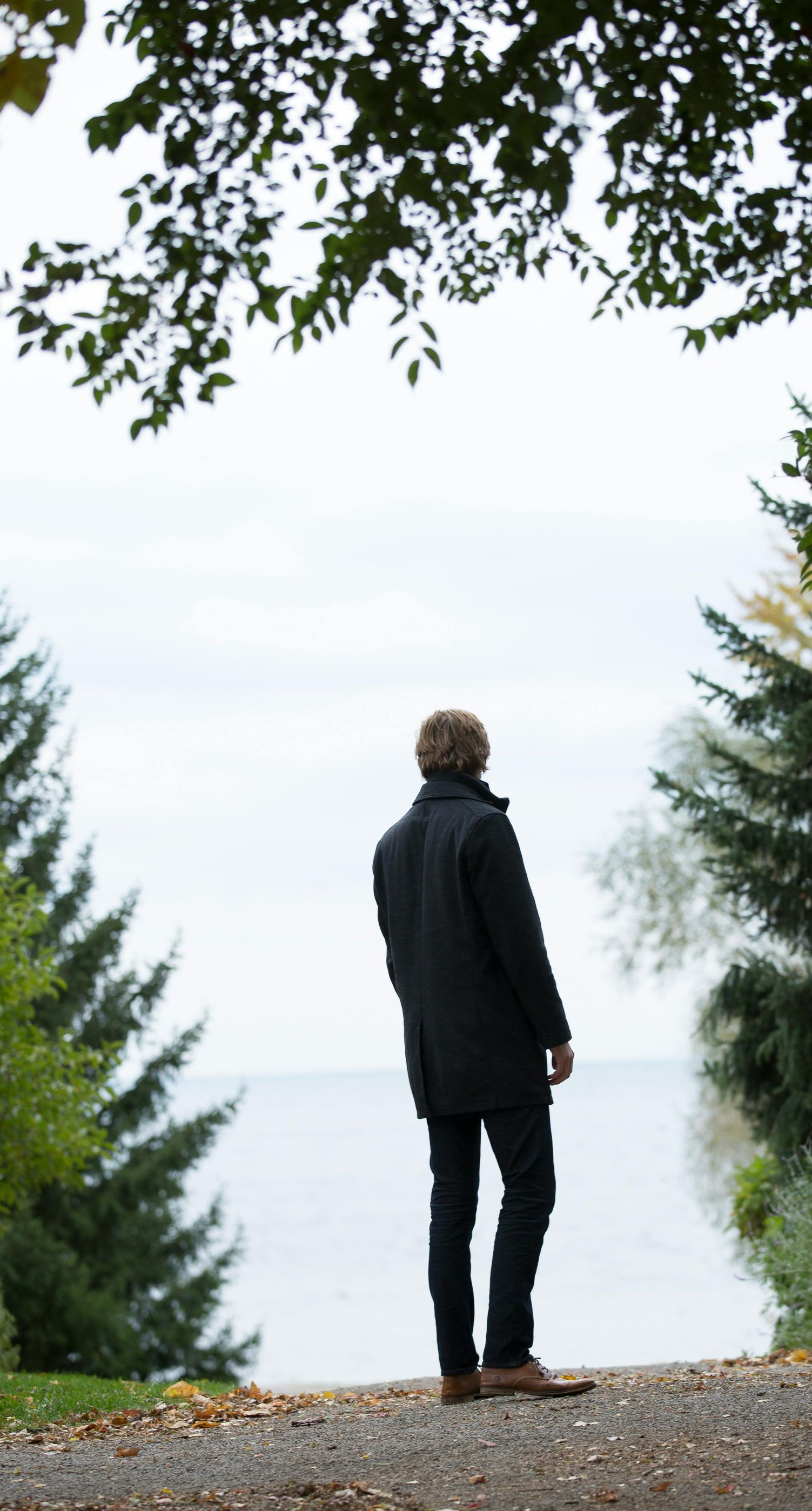 Man in black coat standing, facing water, framed by trees. Overcast sky.