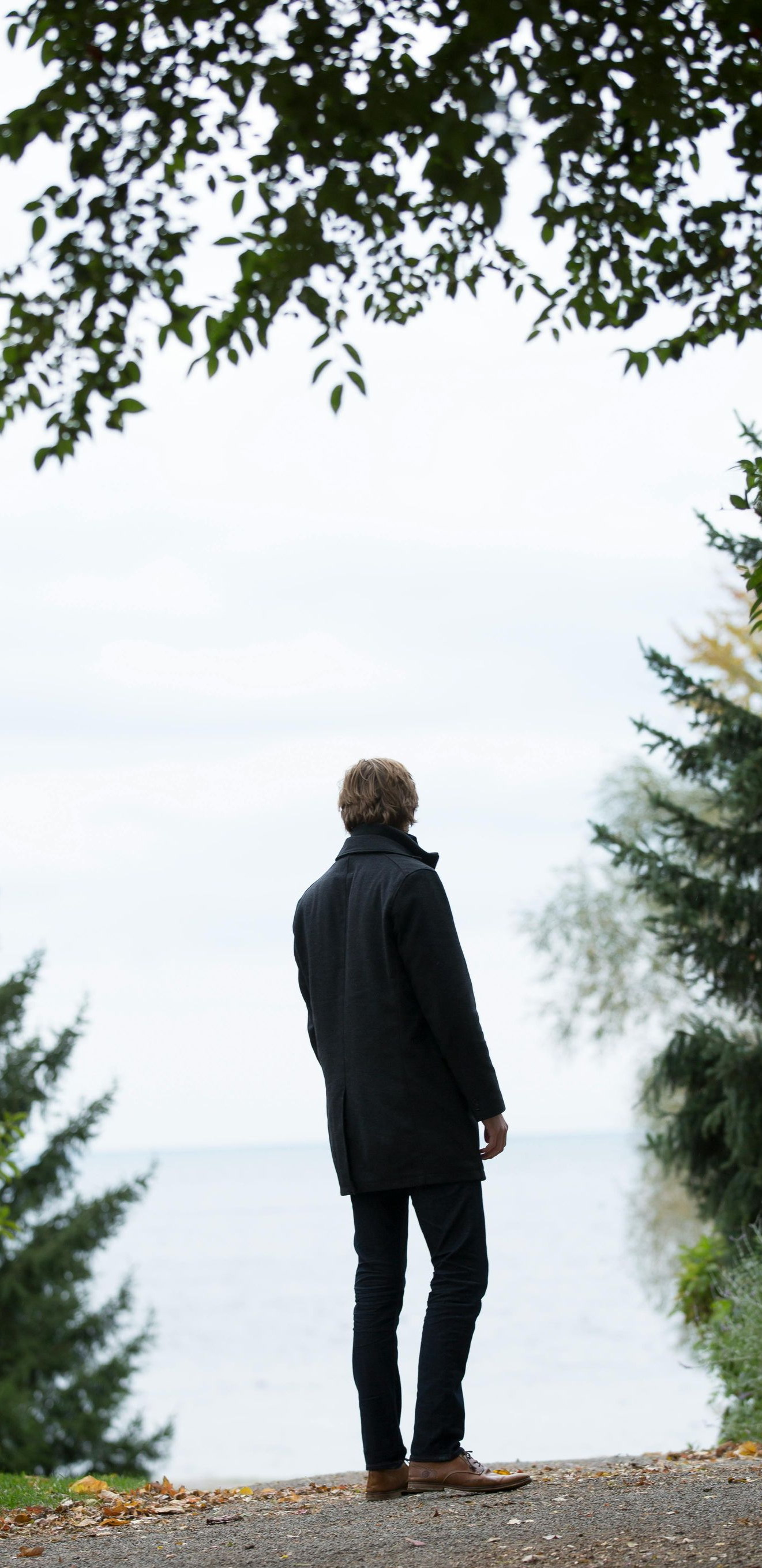 Man in black coat standing, facing water, framed by trees. Overcast sky.