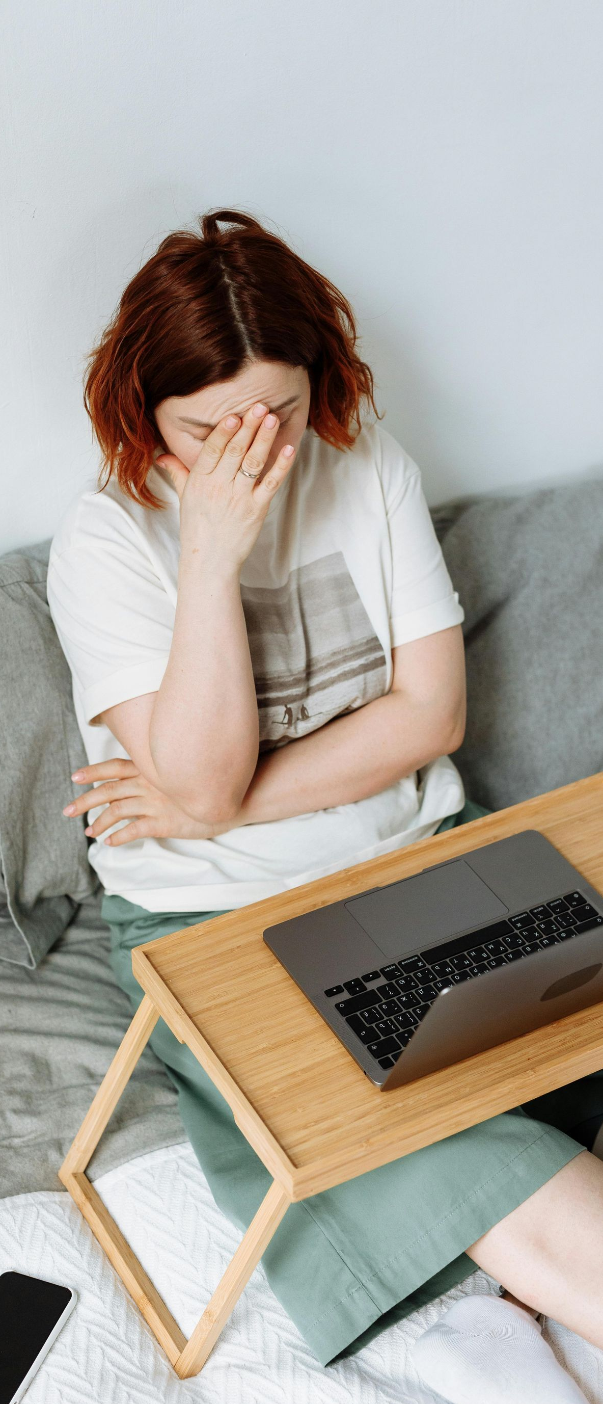 Person with red hair, stressed, face in hand, sitting on bed with laptop on lap desk.
