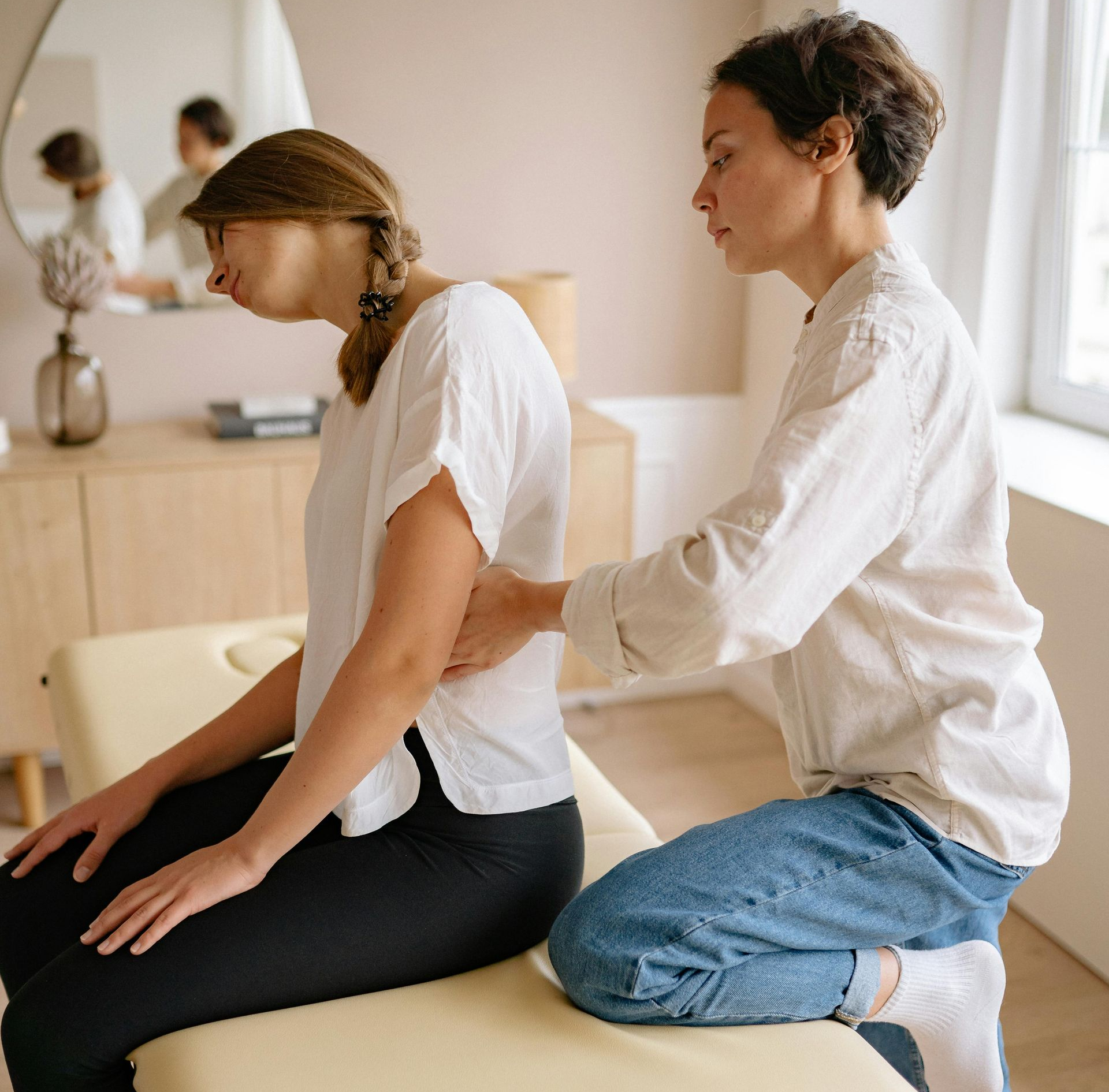Woman receiving back treatment from another woman. Indoors; white shirt, jeans.