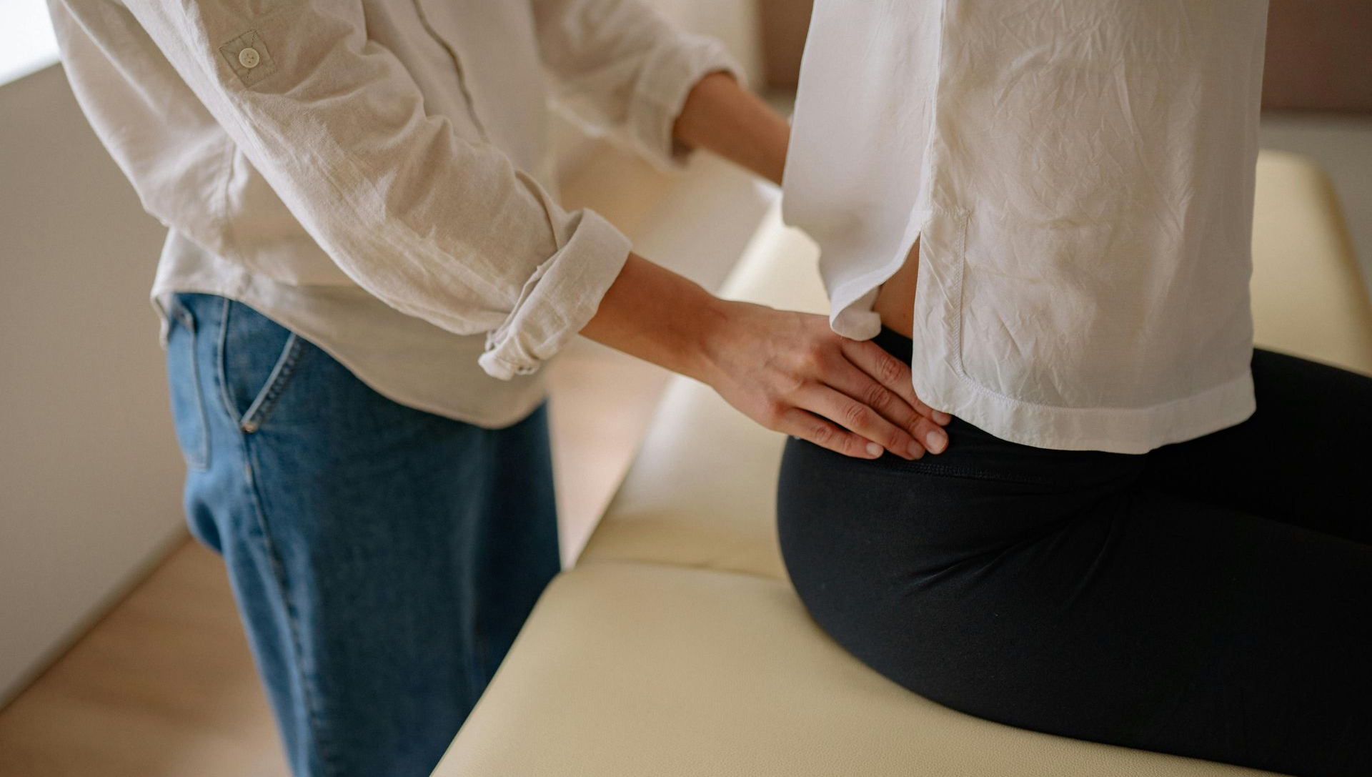 Person's hands on another's lower back, possibly providing treatment. Both are wearing white shirts, one in jeans.