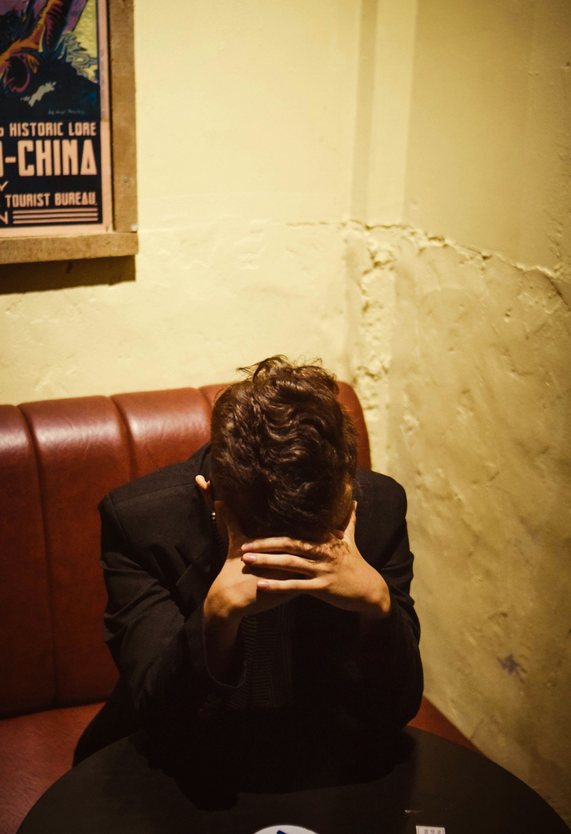 Man with head in hands at a table, in a booth with red leather seating. Low-lit interior, vintage poster.