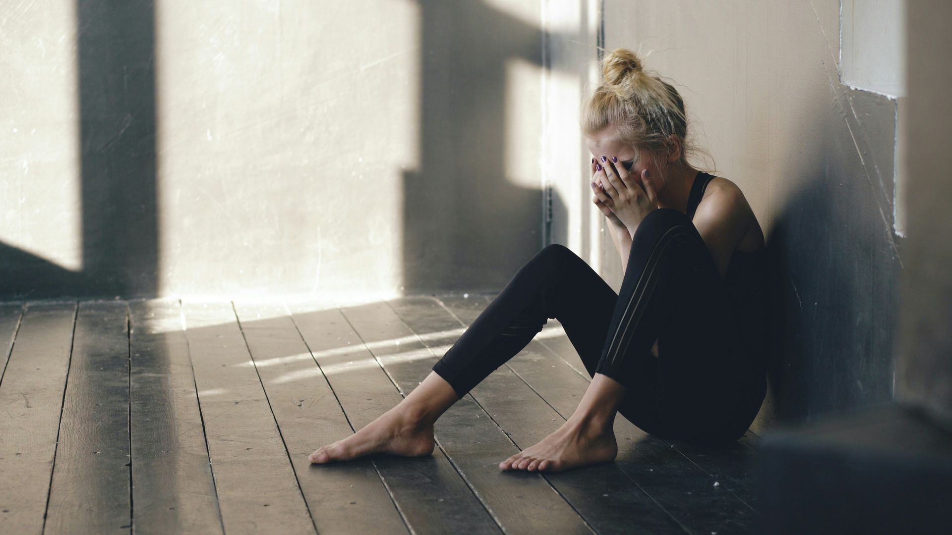 Woman sitting on floor, head in hands, appears distressed. Black athletic wear, light-filled room.