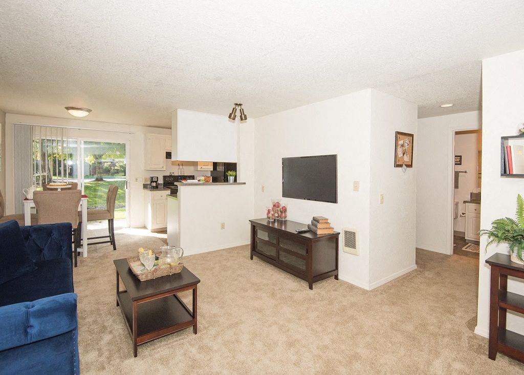 Living room with beige carpet, white walls, and a TV console, open to a small kitchen and dining area.