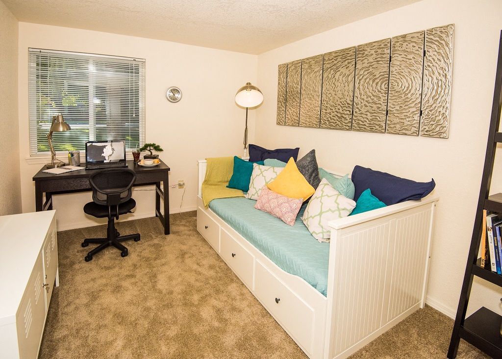 Small apartment bedroom with a white daybed, dark desk by the window, swivel chair, and a tall bookshelf.