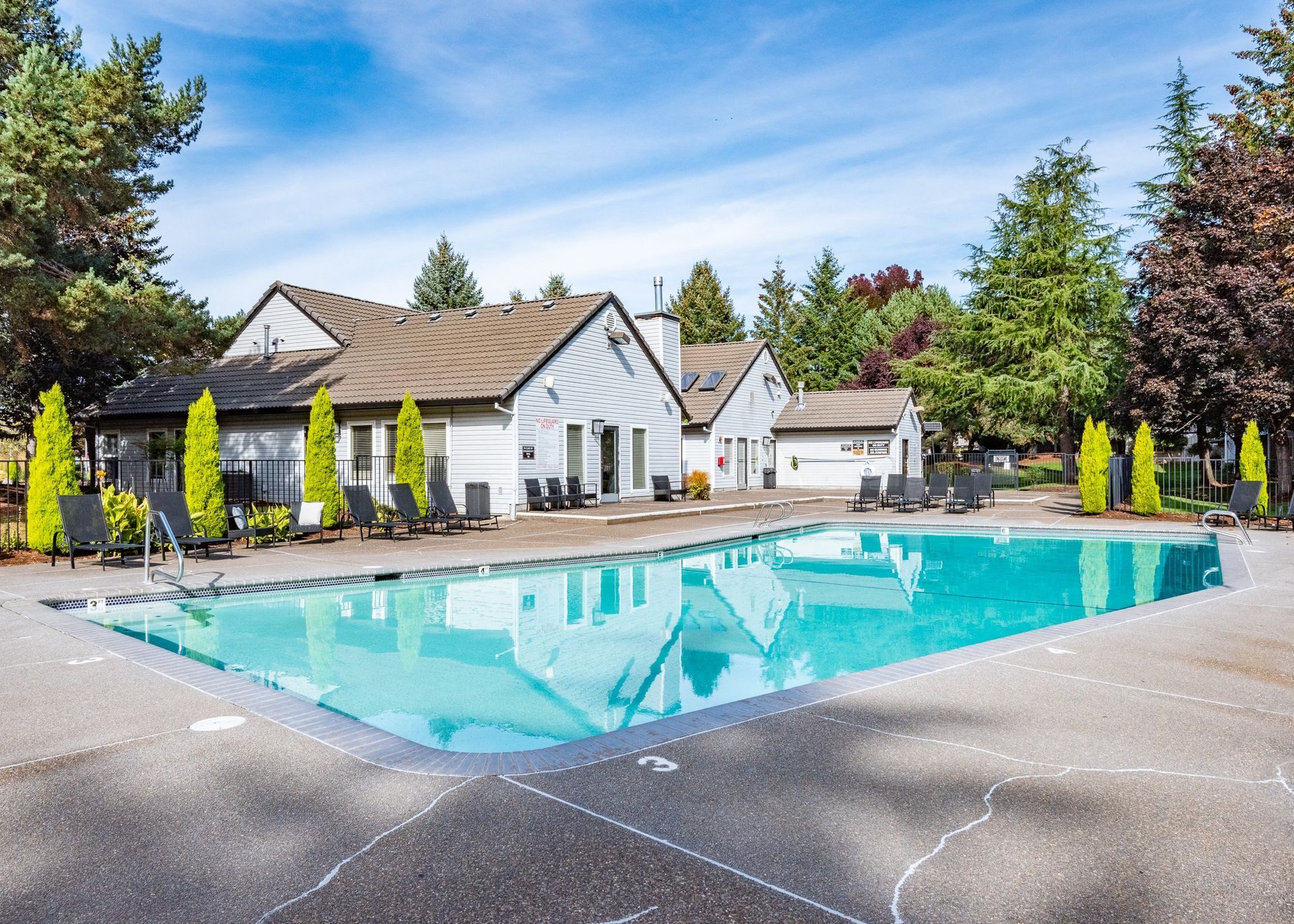 Outdoor community pool with lounge chairs and light-gray buildings, trees in the background.