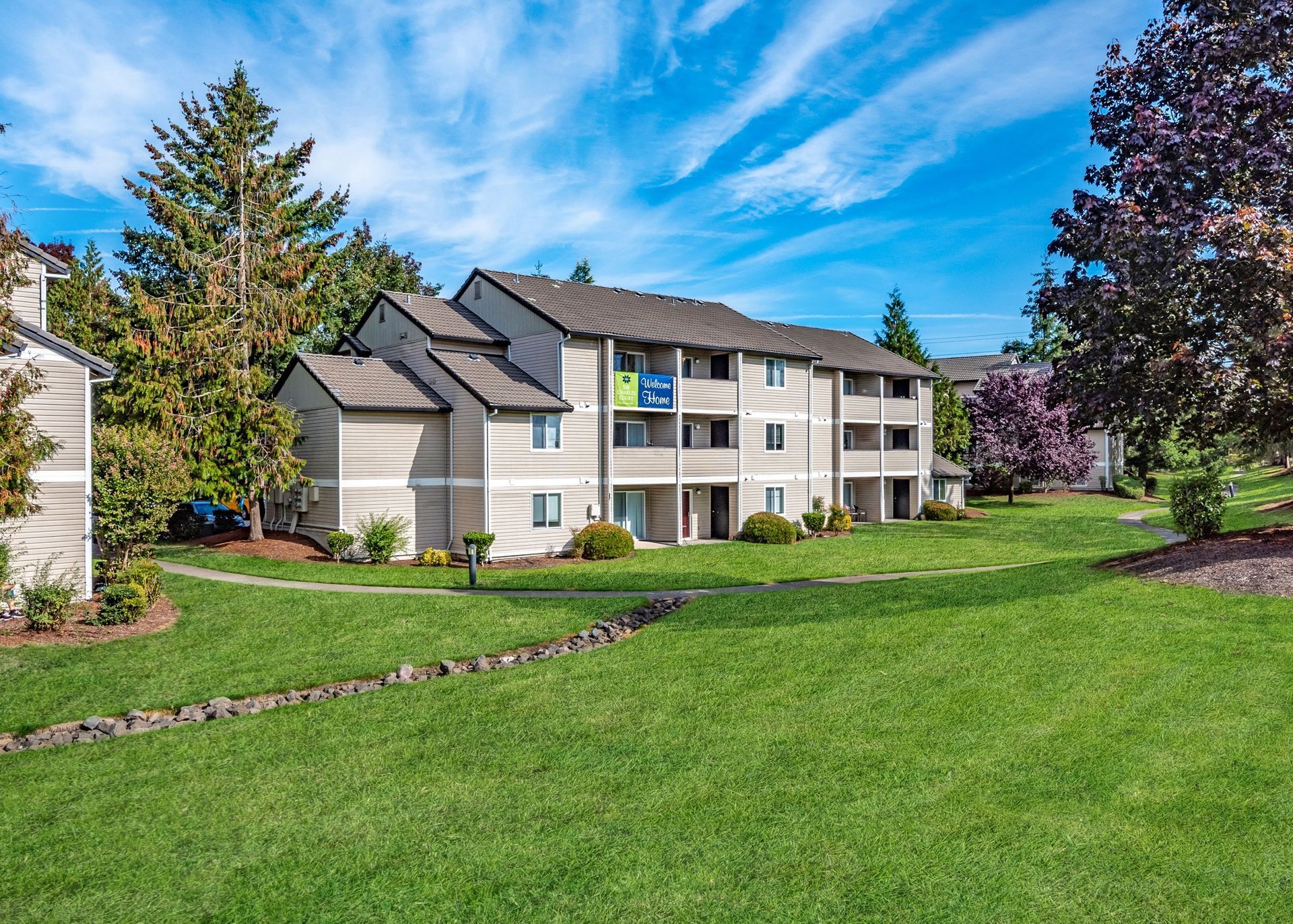 Exterior view of a beige apartment building complex with a green lawn and trees.