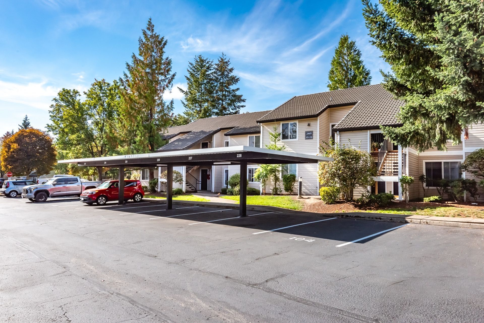 Exterior view of a multi-family apartment community with a long carport and parked cars under a blue sky.