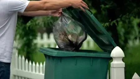 A man is putting a bag of trash into a green trash can.