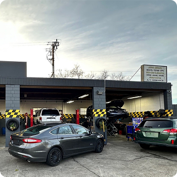Cars parked under a covered auto shop bay, with one lifted and mechanics working inside. | Costa Auto Care and TIre