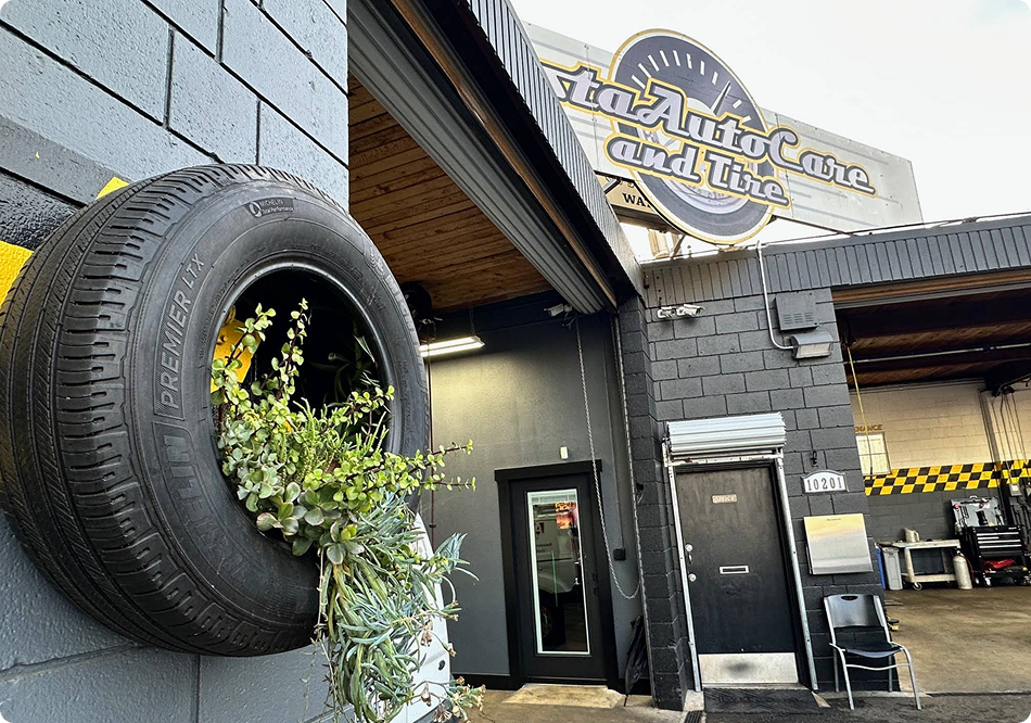 Exterior of a rustic tattoo shop with a tire planter, hanging sign, and black storefront. | Costa Auto Care and TIre