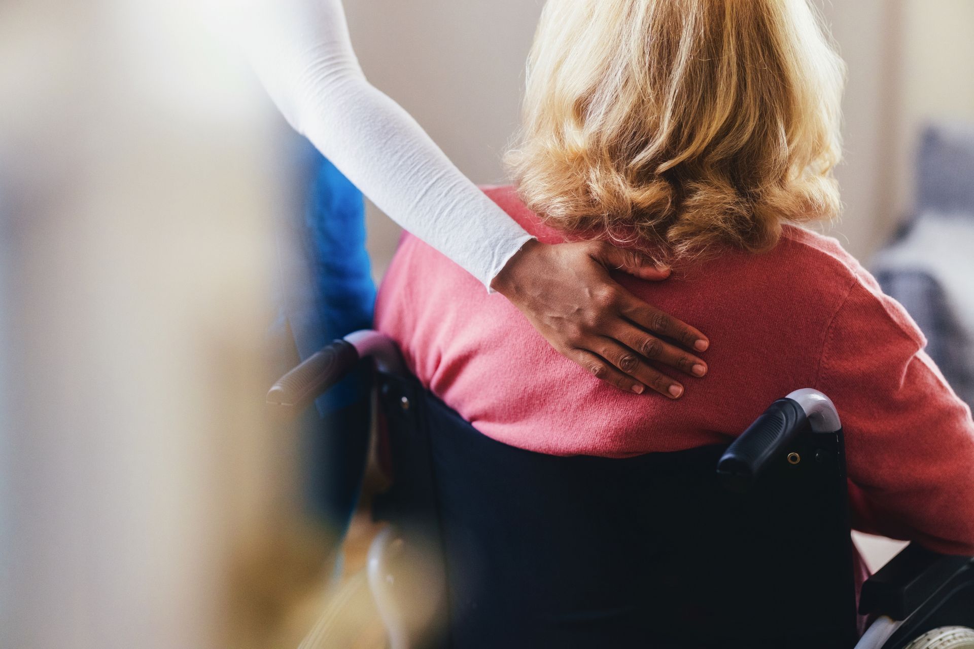 Person's hand on the back of a woman in a wheelchair; indoors.
