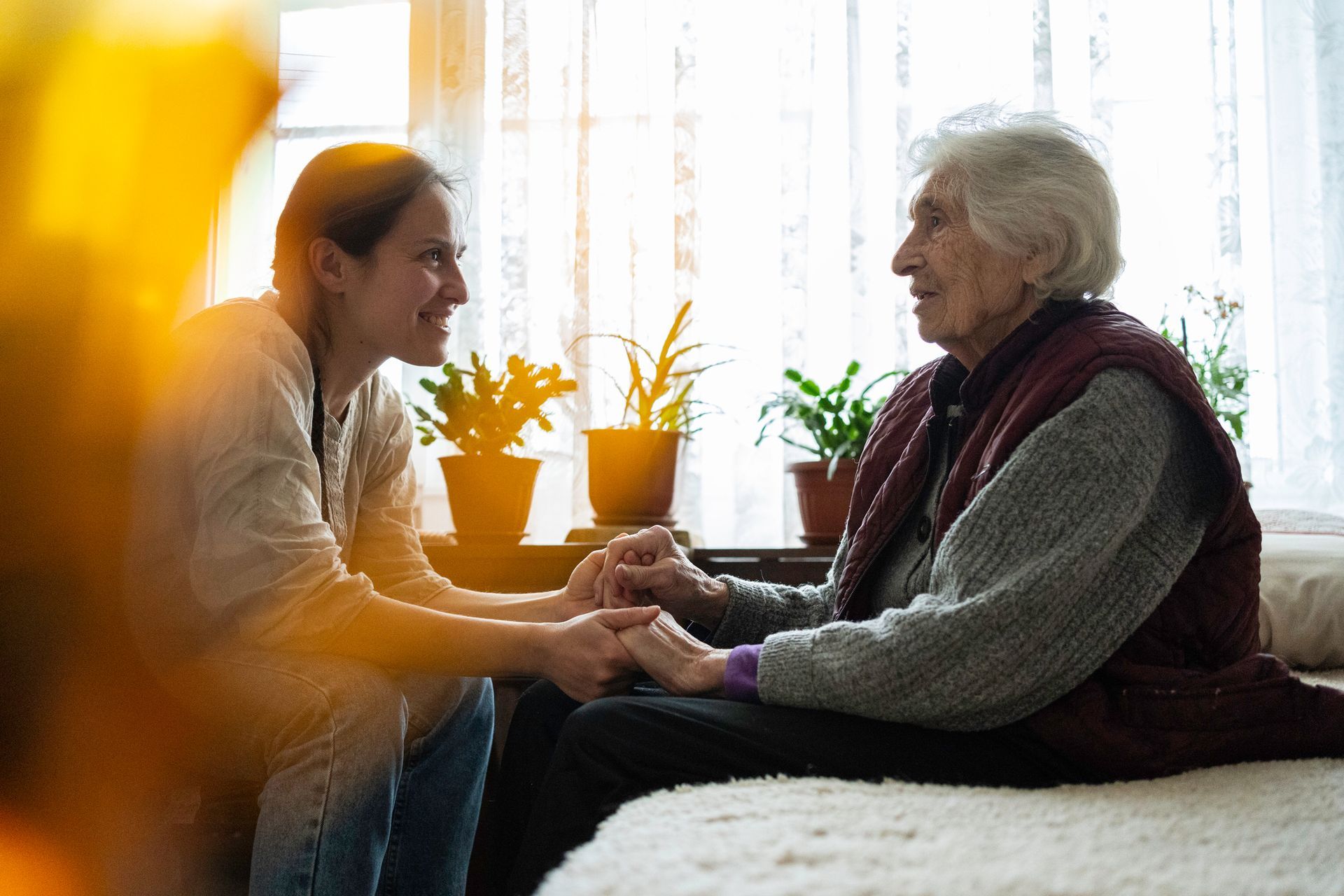 A woman is holding the hand of an elderly woman while sitting on a bed.